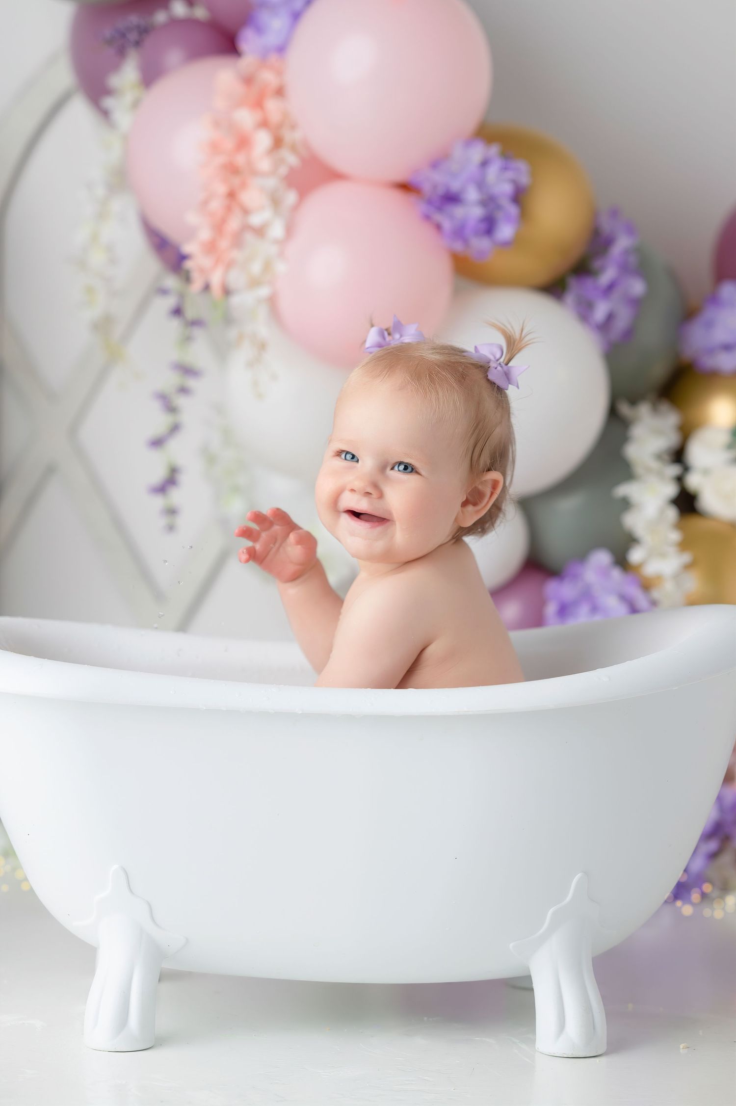 Smiling baby girl sitting in a white clawfoot bathtub surrounded by pink, purple, and gold balloons and flowers.