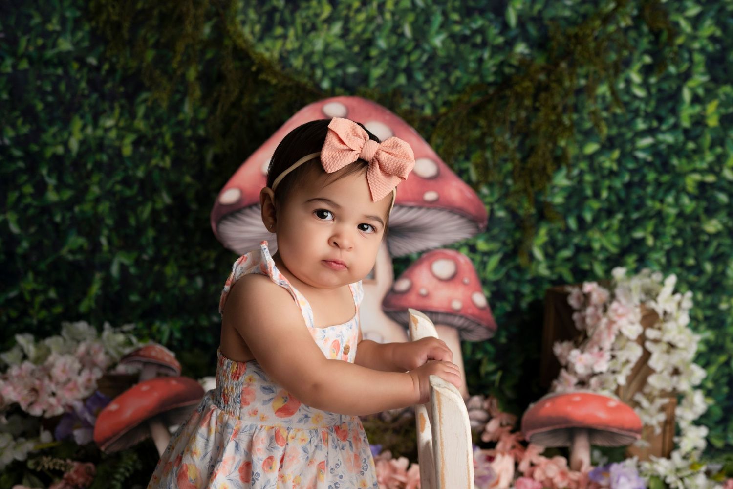 A baby in a pink dress and bow sits among pink flowers in a garden setting for a professional photo shoot.