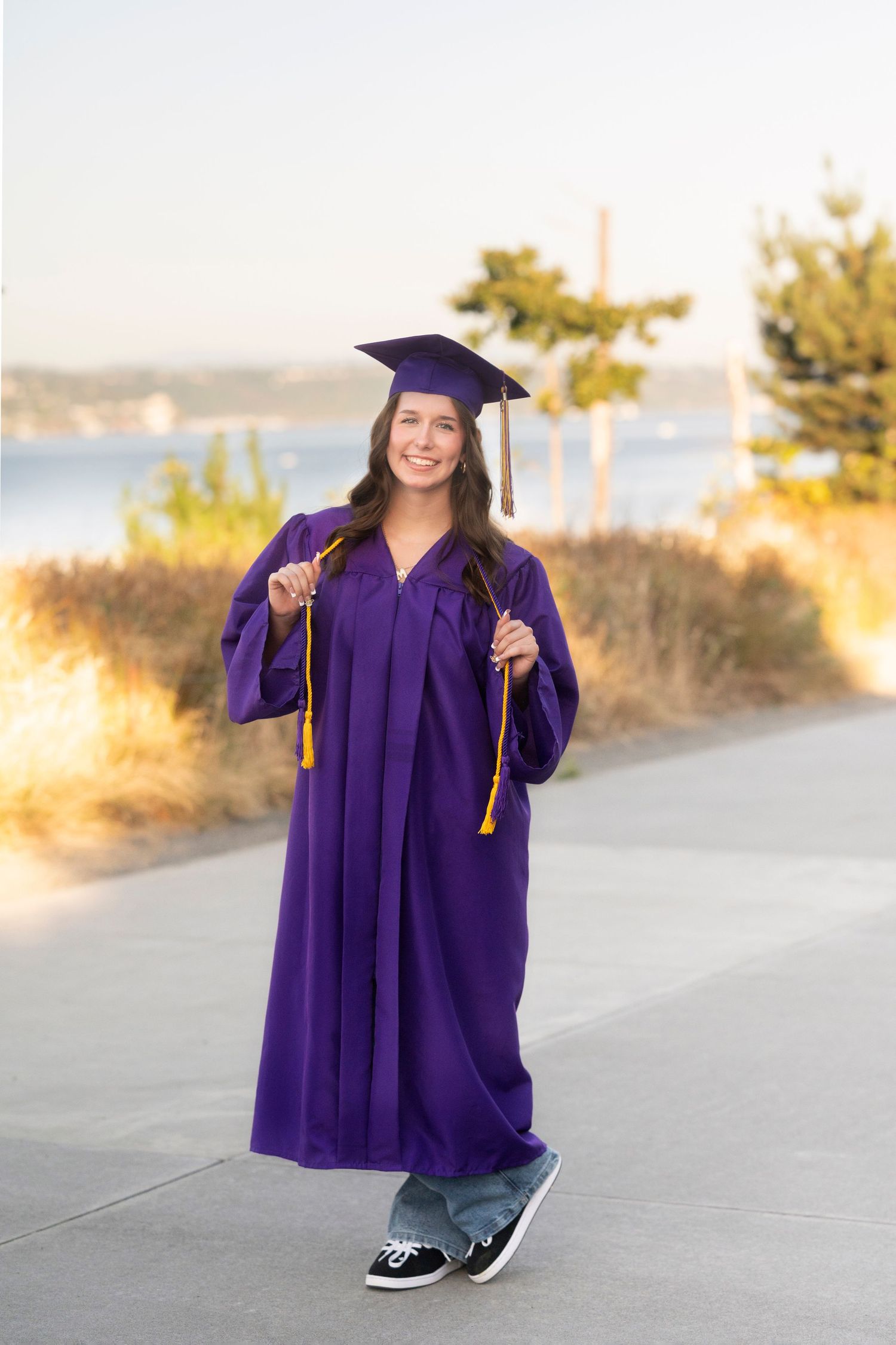 Graduate in purple cap and gown poses outdoors during sunset for graduation photos on a coastal path.