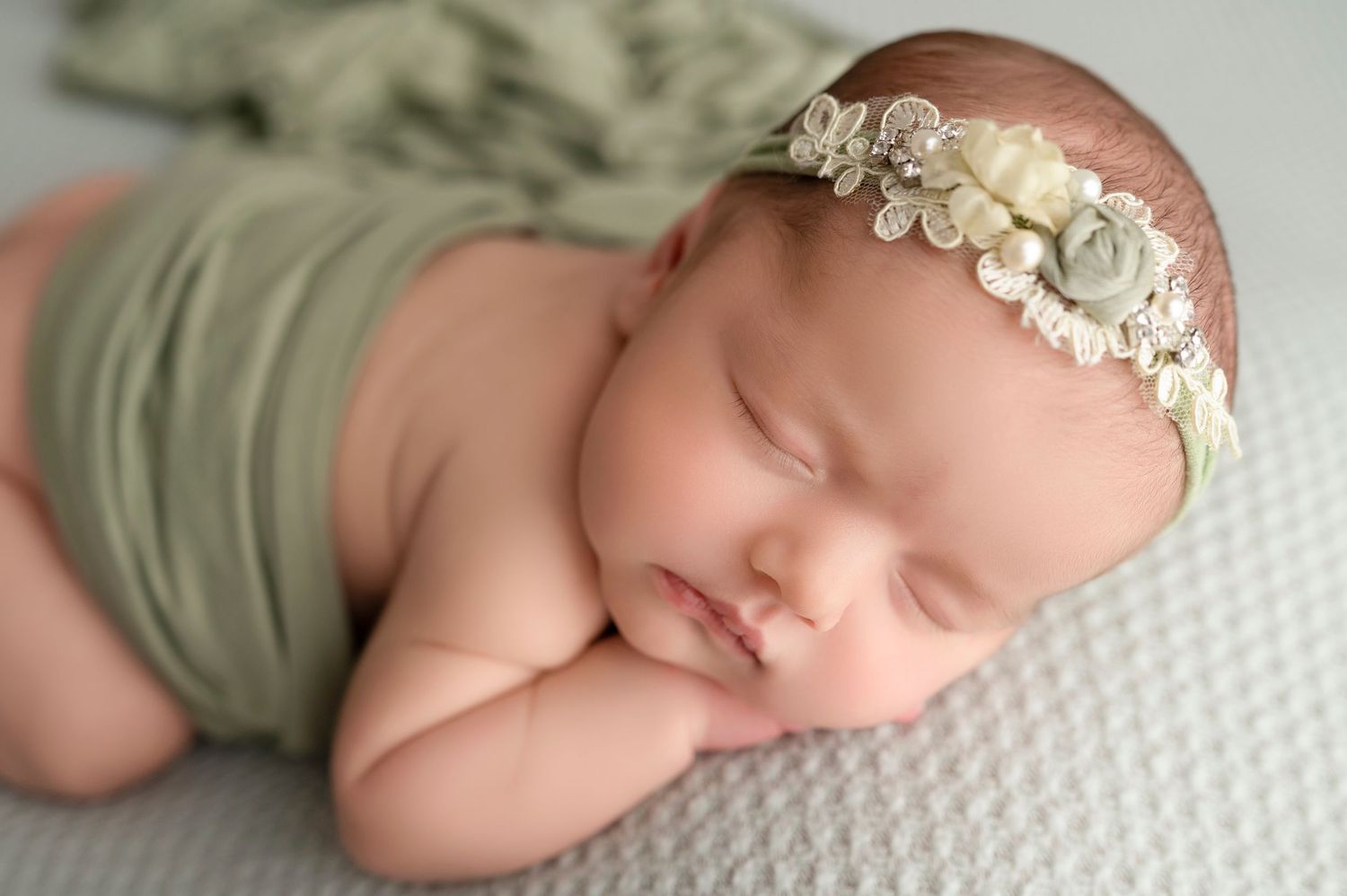 Peaceful newborn in sage green wrap and floral headband sleeps on textured surface.