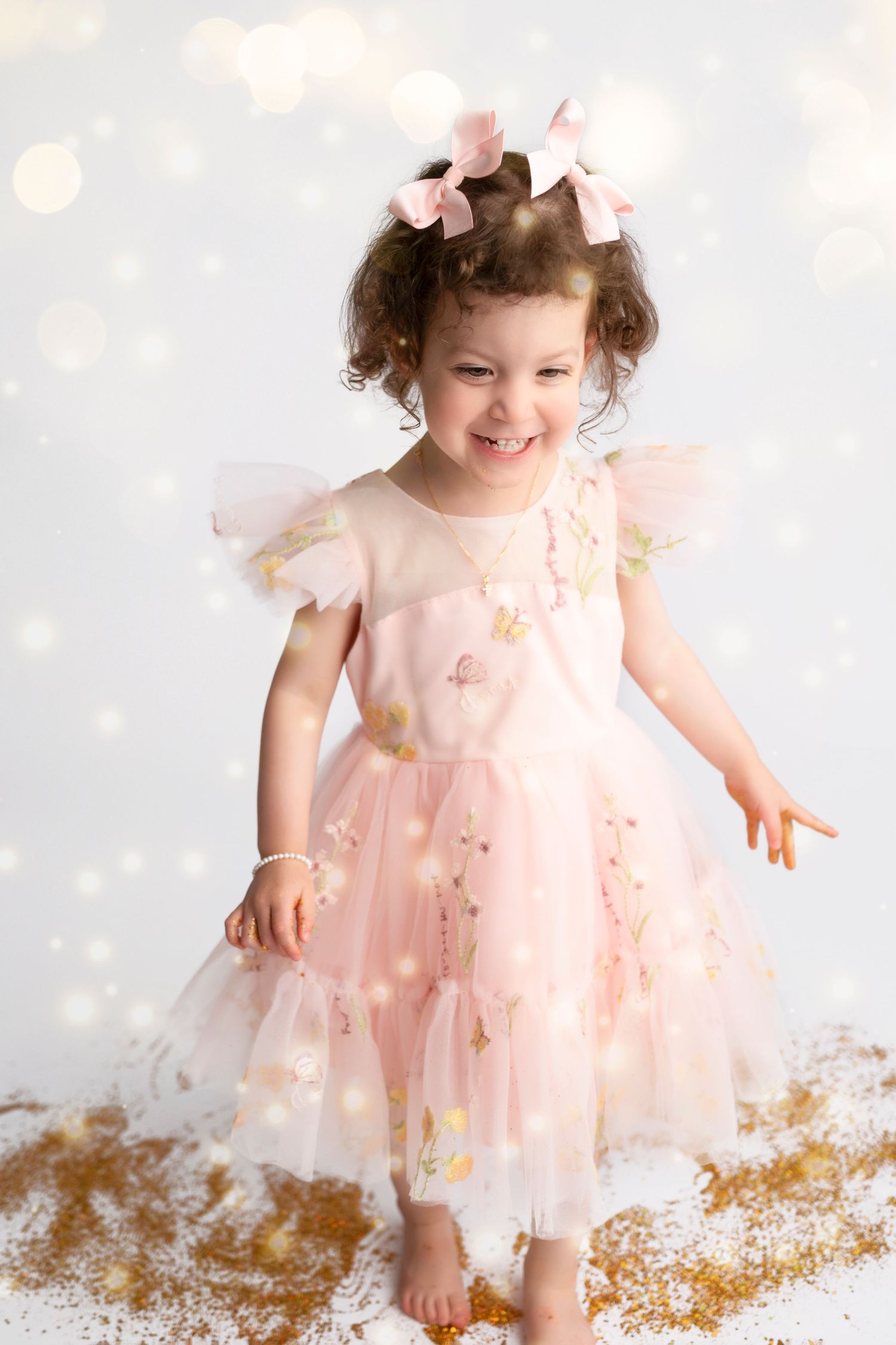 A small child in a pink tulle dress plays with gold glitter on a white background with bokeh lights.