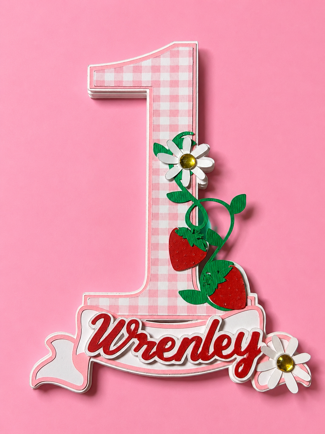 Pink gingham number 1 cake topper with strawberries, daisies, and the name Wrenley on a banner.