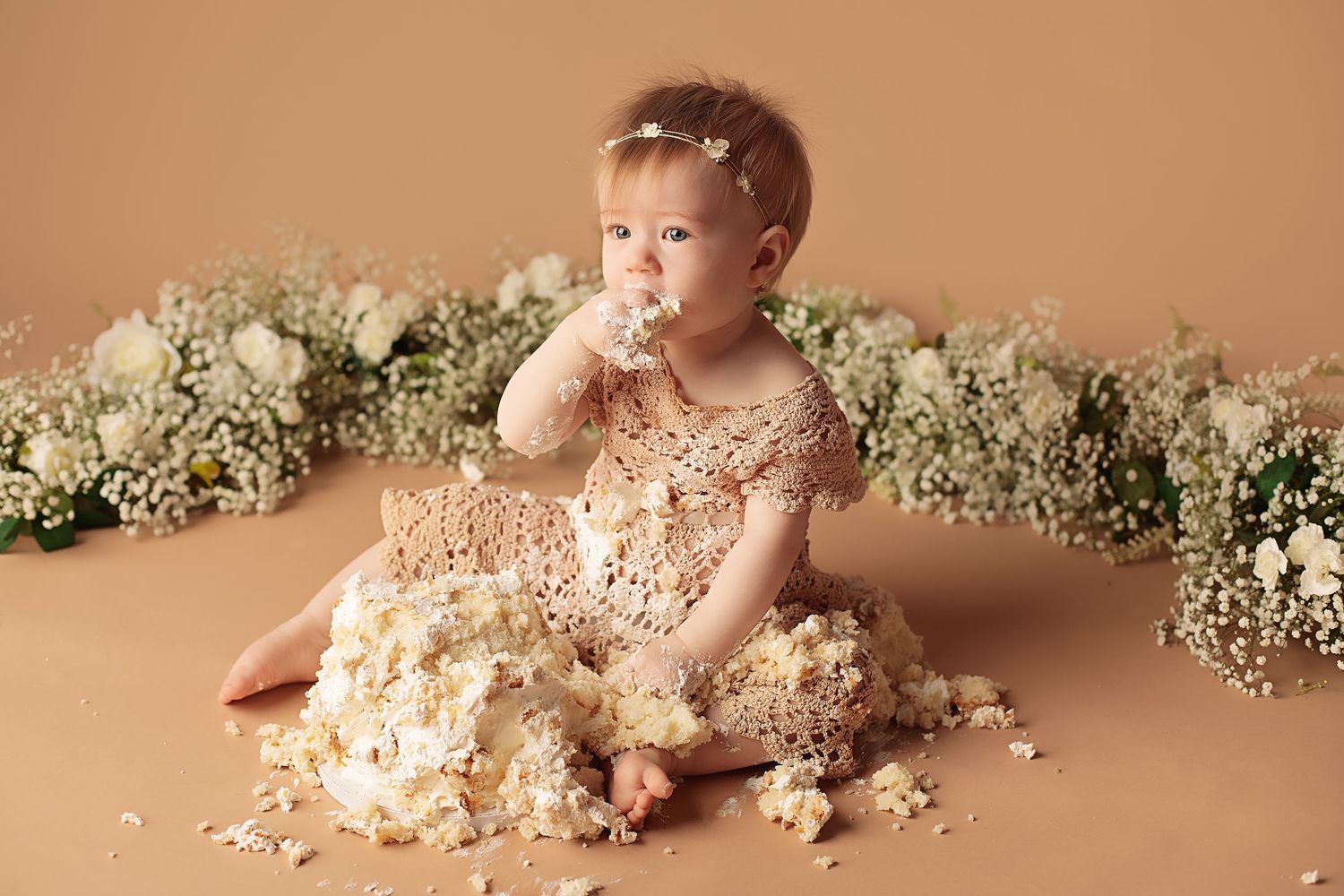 Baby in white lace outfit dives face-first into cake during first birthday photoshoot with floral decorations.