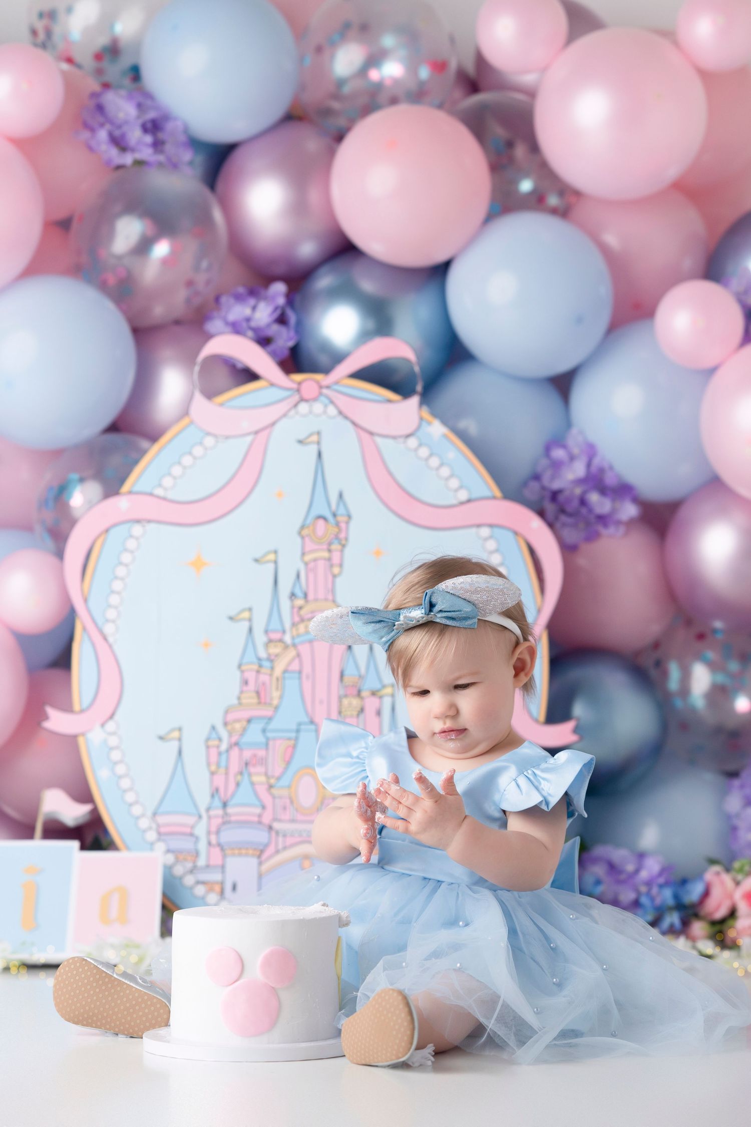 Baby girl in blue dress at Cinderella castle first birthday party with pink and purple balloon backdrop.