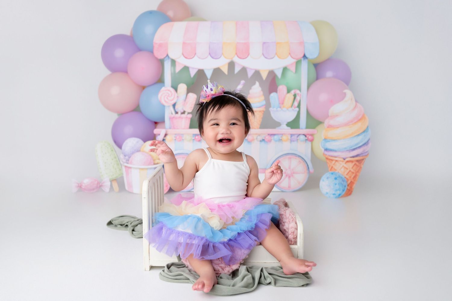 A joyful baby girl in a rainbow tutu sits smiling at a pastel ice cream shop birthday cake smash setup.