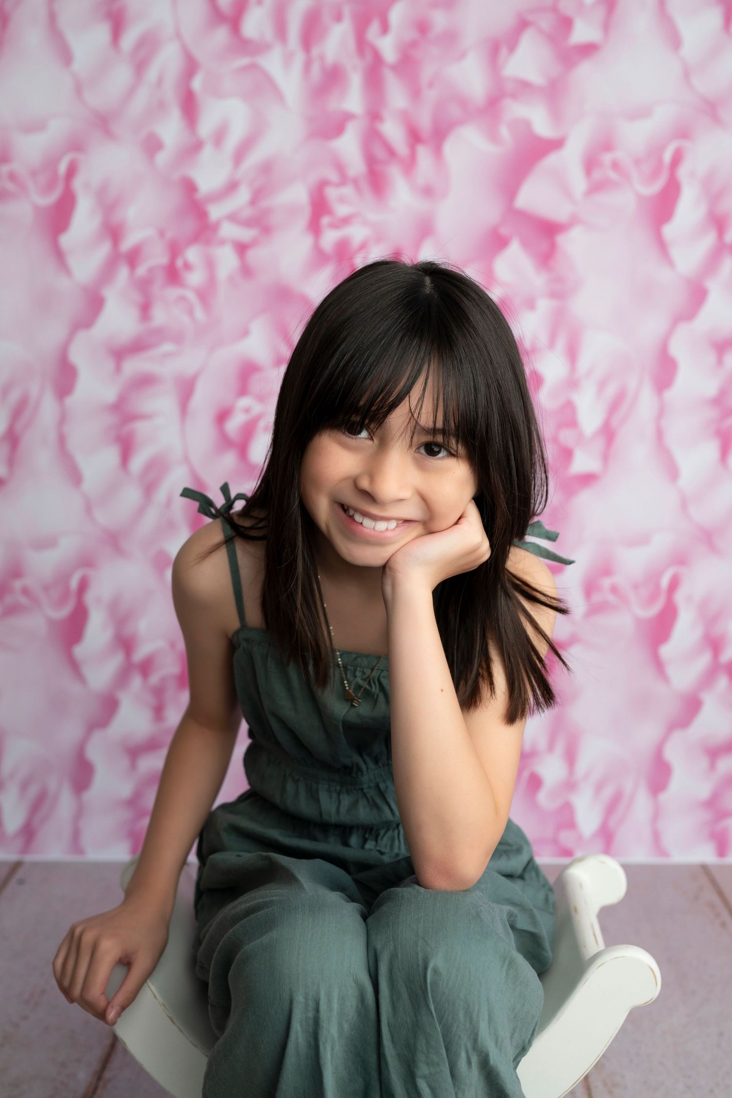 A smiling young girl in a green jumpsuit sits on a white chair against a pink floral backdrop.