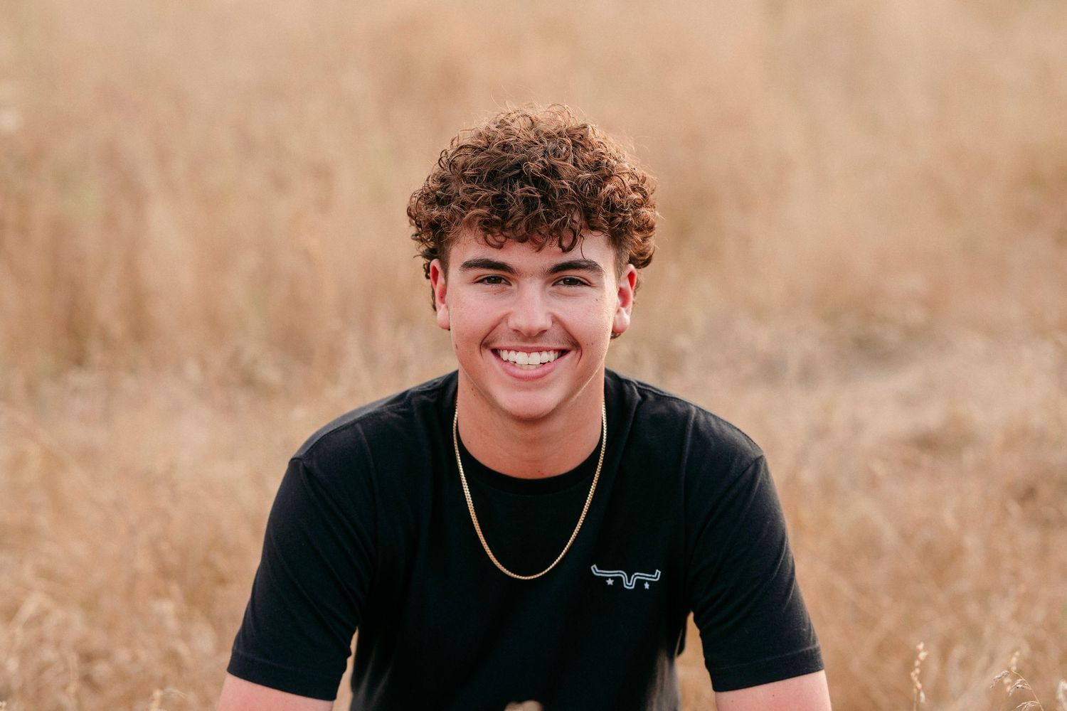Person wearing black t-shirt and jeans smiles while standing in a field with golden autumn colors behind.