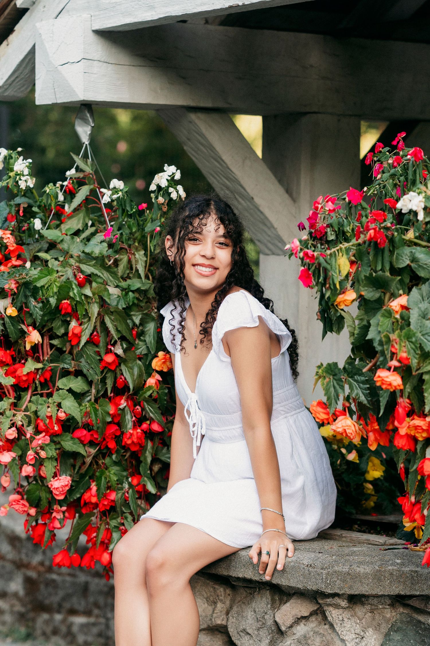 A woman in a white dress poses gracefully among vibrant red and orange flowers in a garden setting.