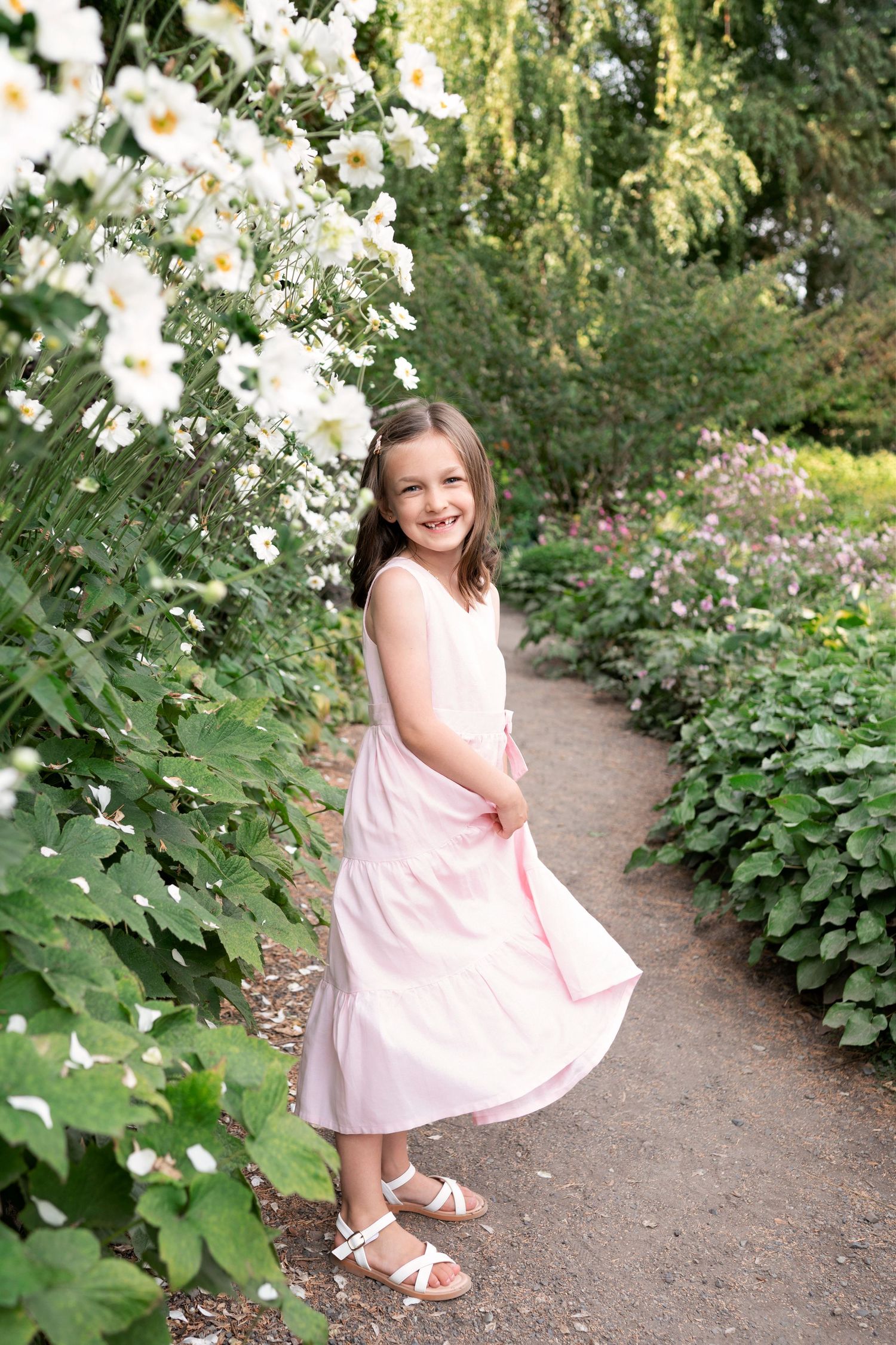 A young figure in a pink summer dress twirls on a garden path lined with white daisies and lush greenery.