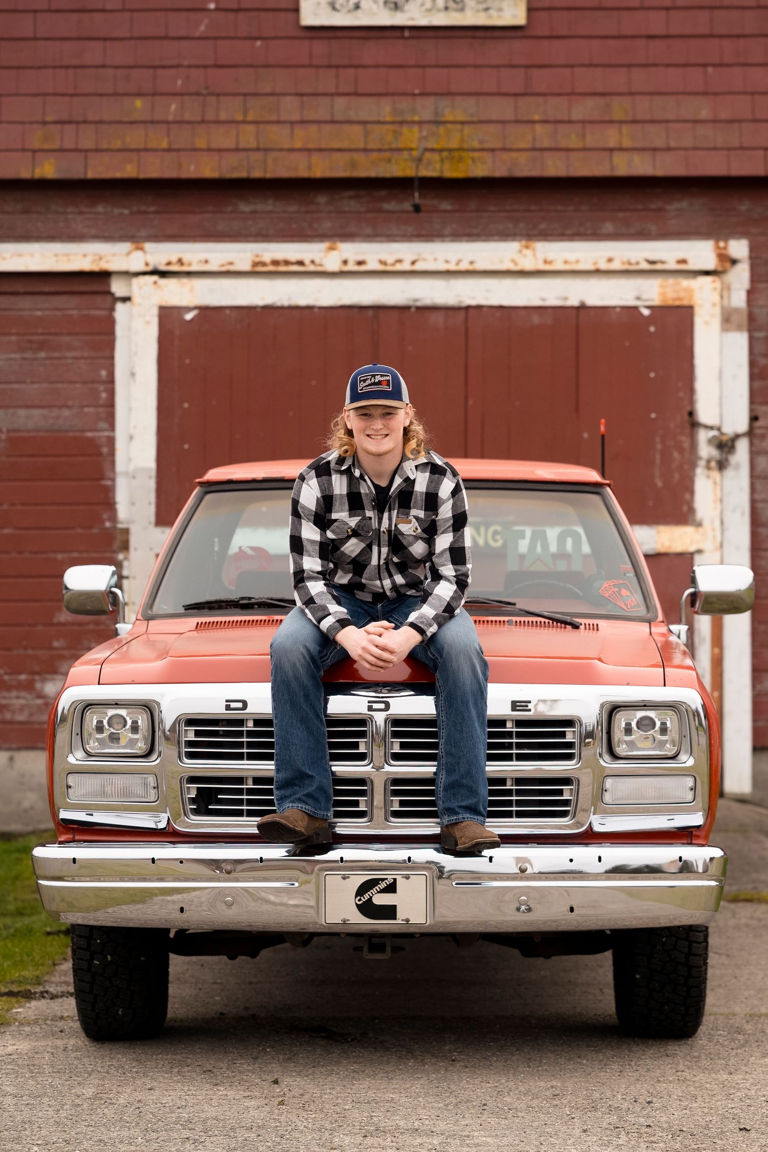 A person in plaid flannel and jeans sits on the hood of a vintage silver car in front of a red barn building.