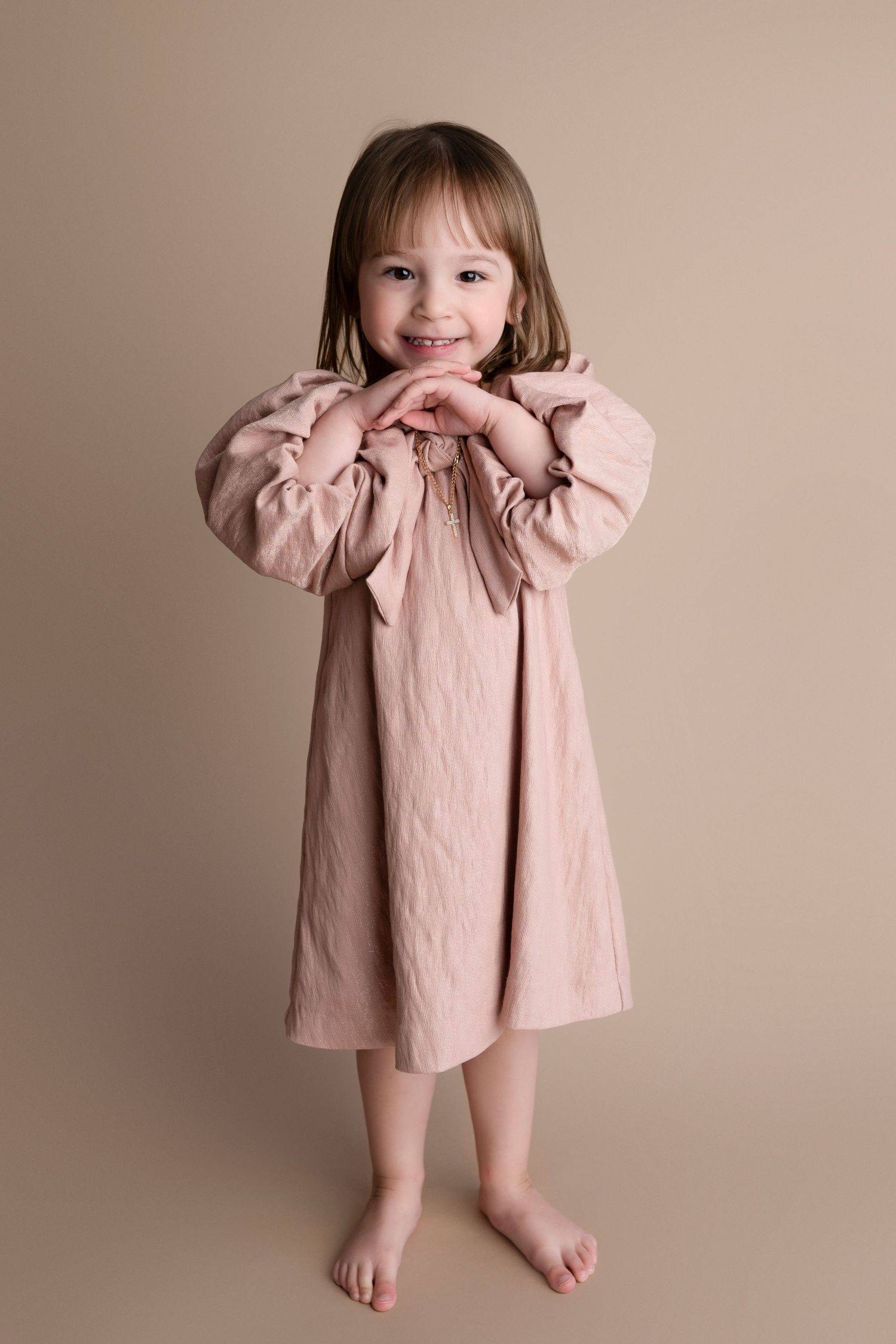 Smiling toddler girl in a pink long-sleeve dress standing barefoot against a beige studio background.