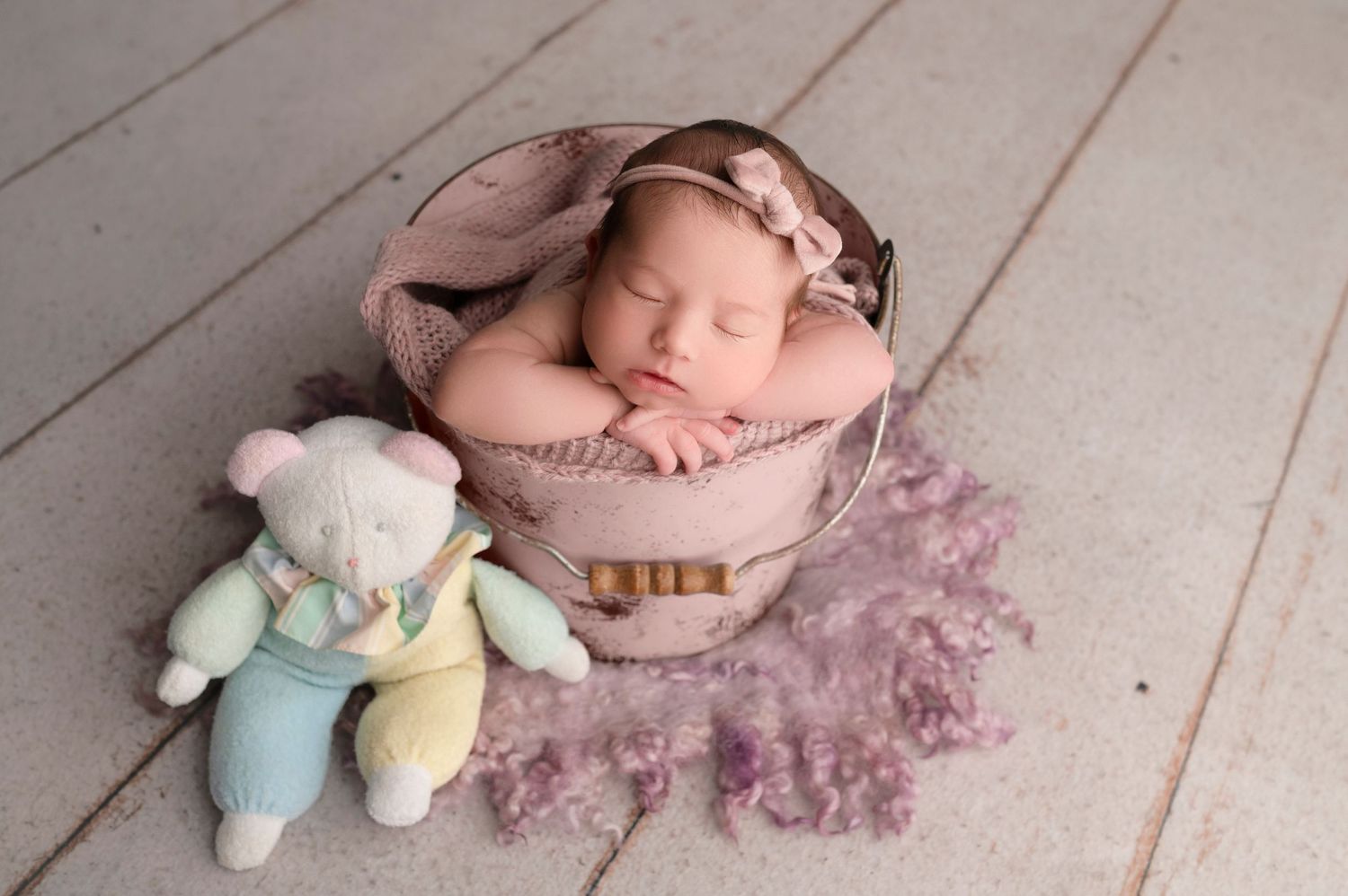 Sleeping newborn baby girl in pink bucket with teddy bear, wearing floral headband, on white wood floor.