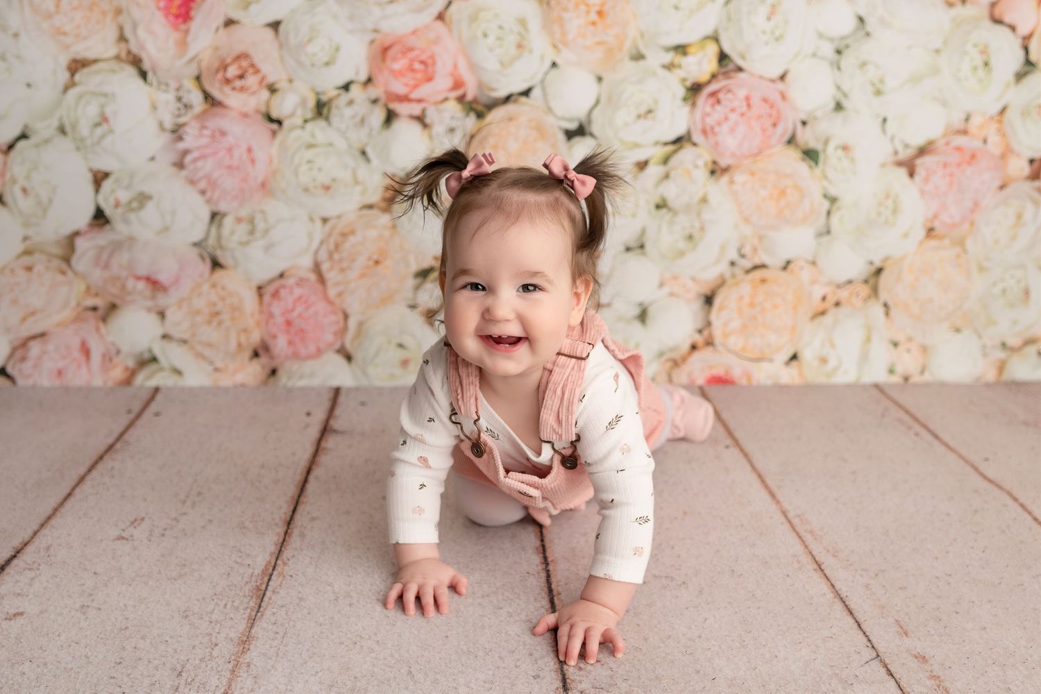 Smiling baby girl in pink outfit crawling on wood floor with floral backdrop during professional photo session.