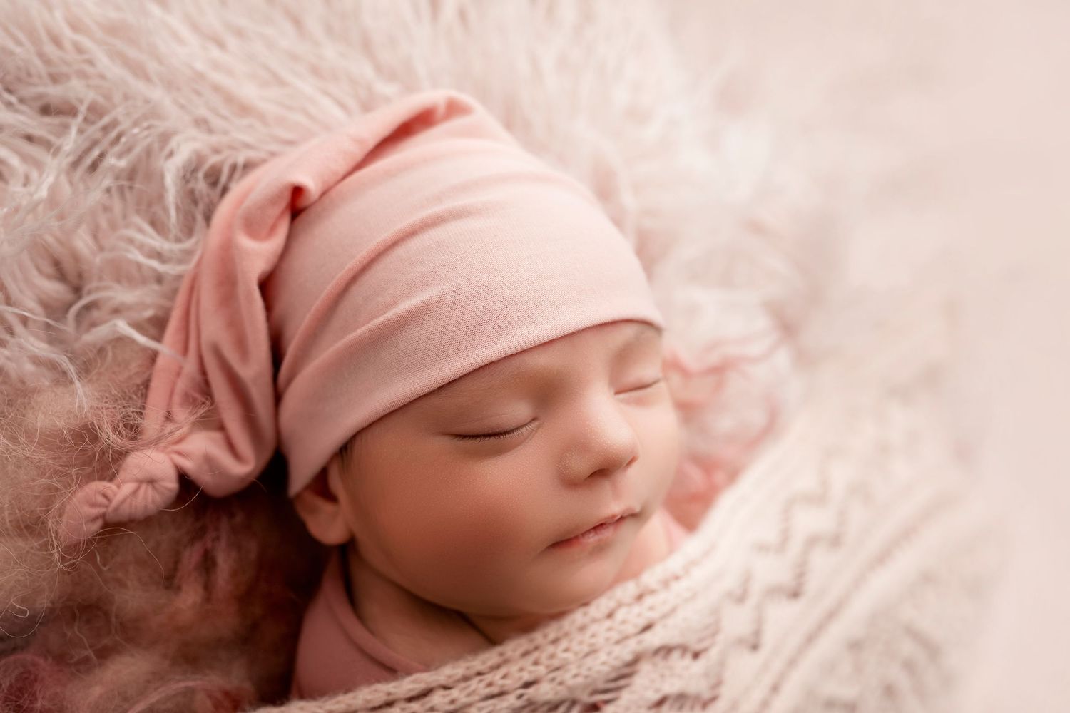 Newborn baby sleeping peacefully in pink swaddle blanket with matching hat against soft textured backdrop.