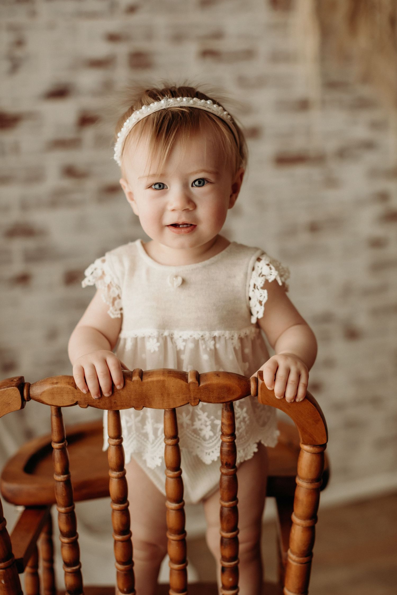 Four sequential photos show a baby sitting at a wooden chair wearing a white dress against a brick background.