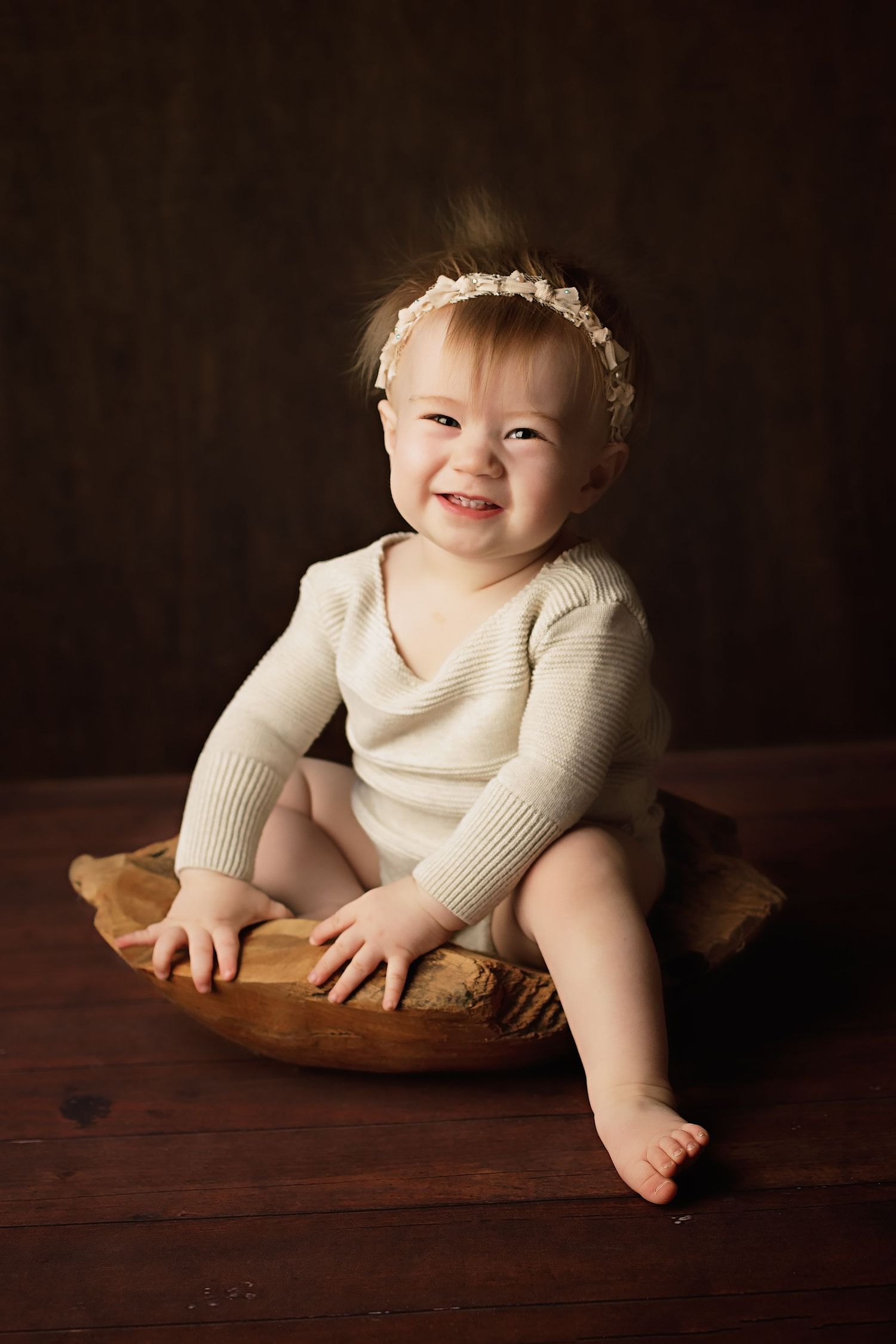 Smiling baby wearing white knit sweater and headband sits in wooden bowl on dark wooden surface.