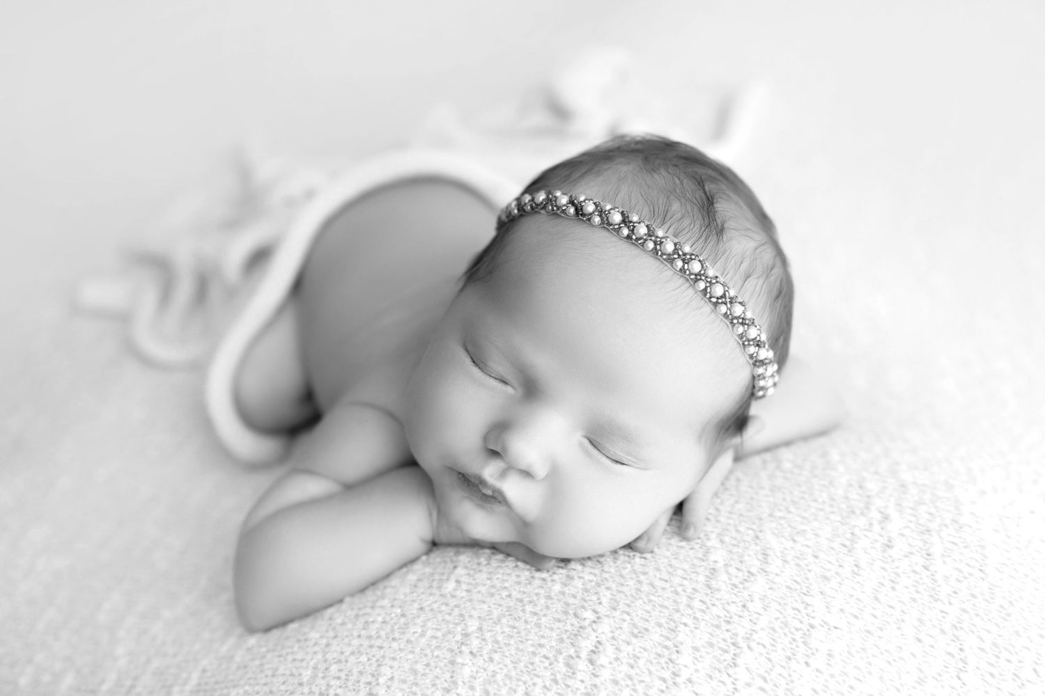 Black and white newborn photo of sleeping baby girl wearing a delicate crystal headband, resting chin on hands.