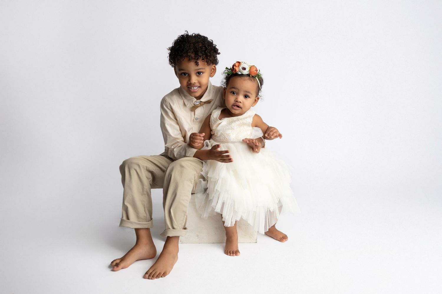Two young children in white and beige formal outfits pose together on a white box against a white background.