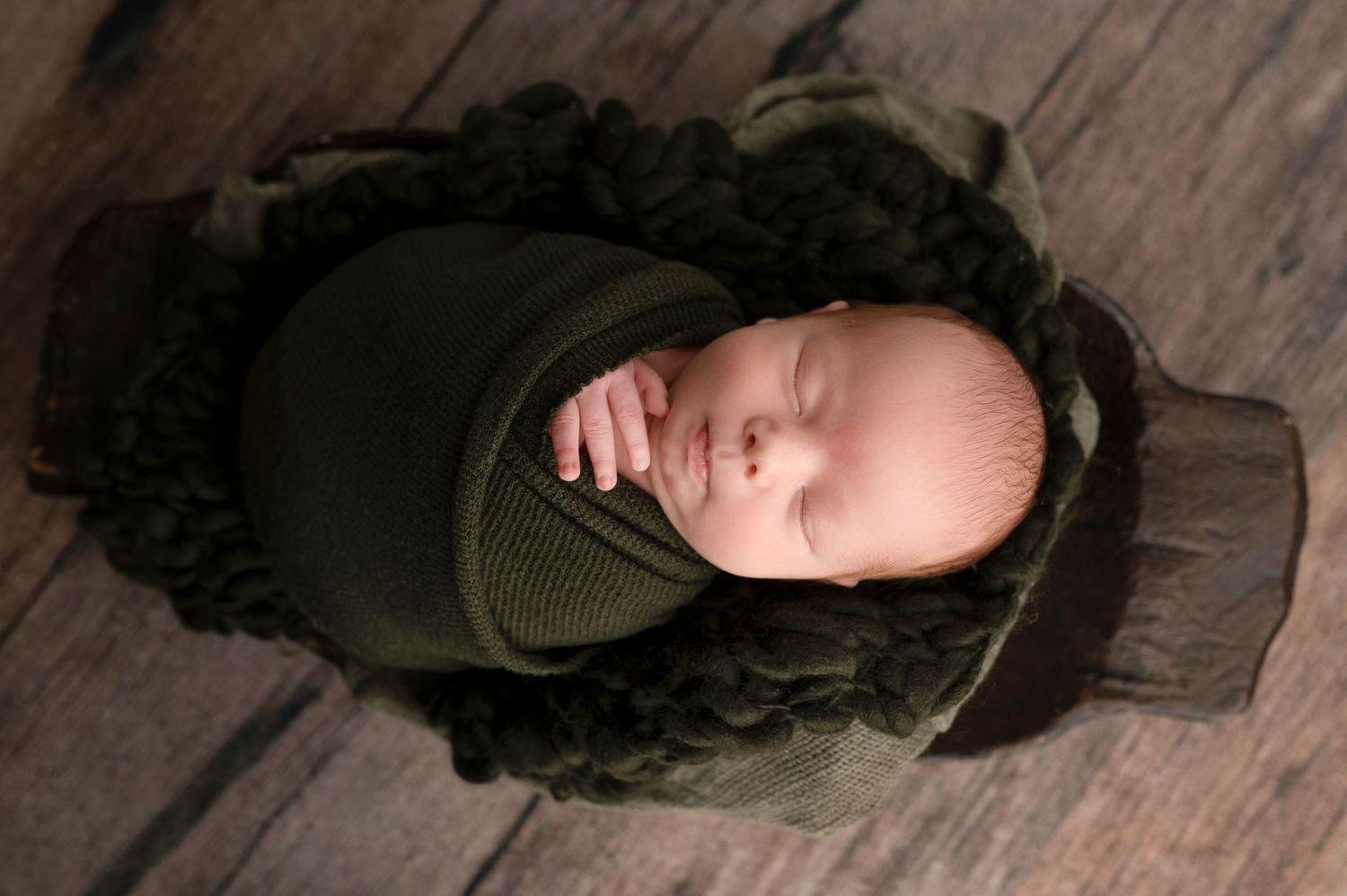 Newborn baby sleeping peacefully wrapped in olive green knit fabric nestled in a dark wooden bowl with fluffy green filler.