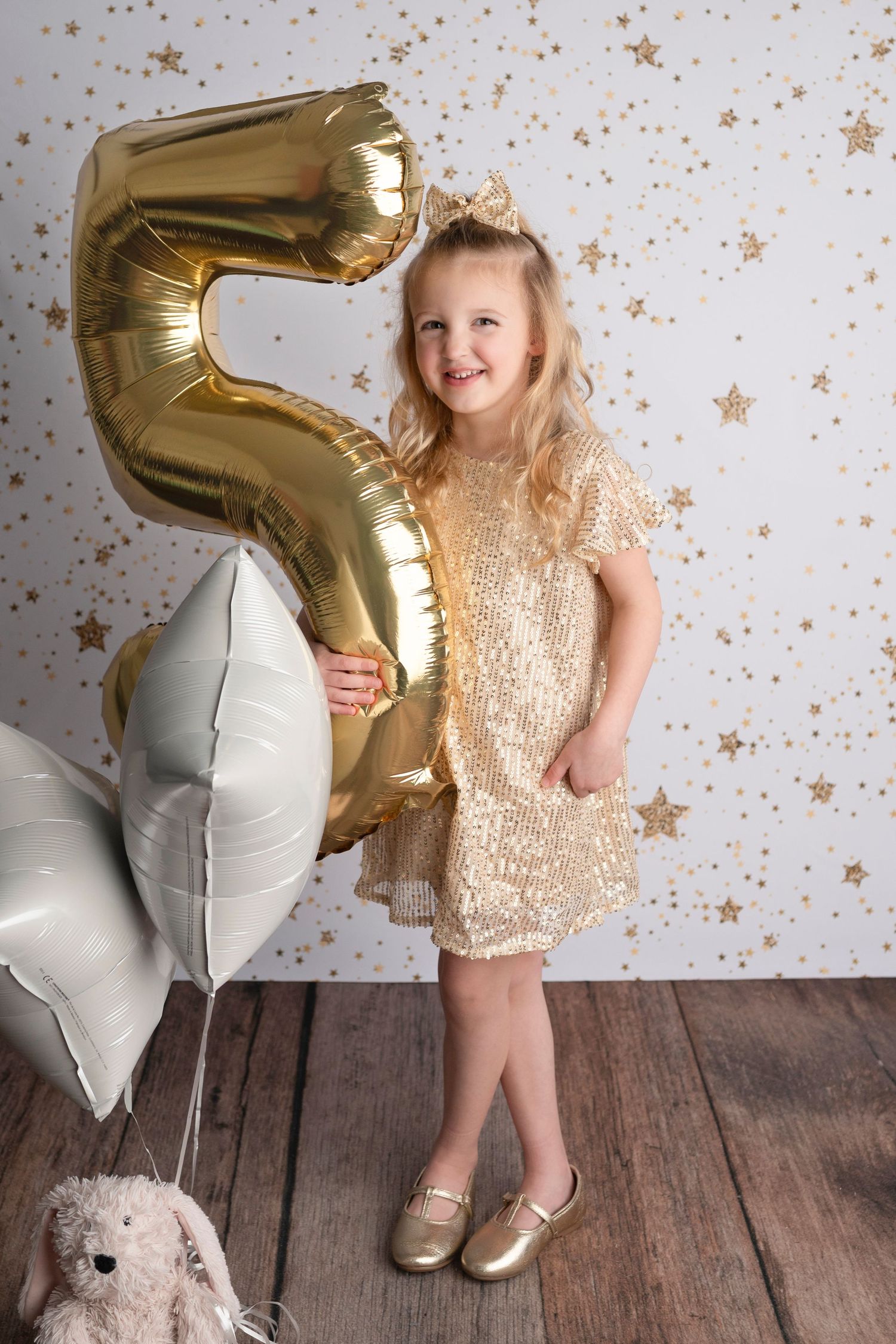 A child in a sparkly gold dress poses with silver and gold number balloons against a star-speckled backdrop.