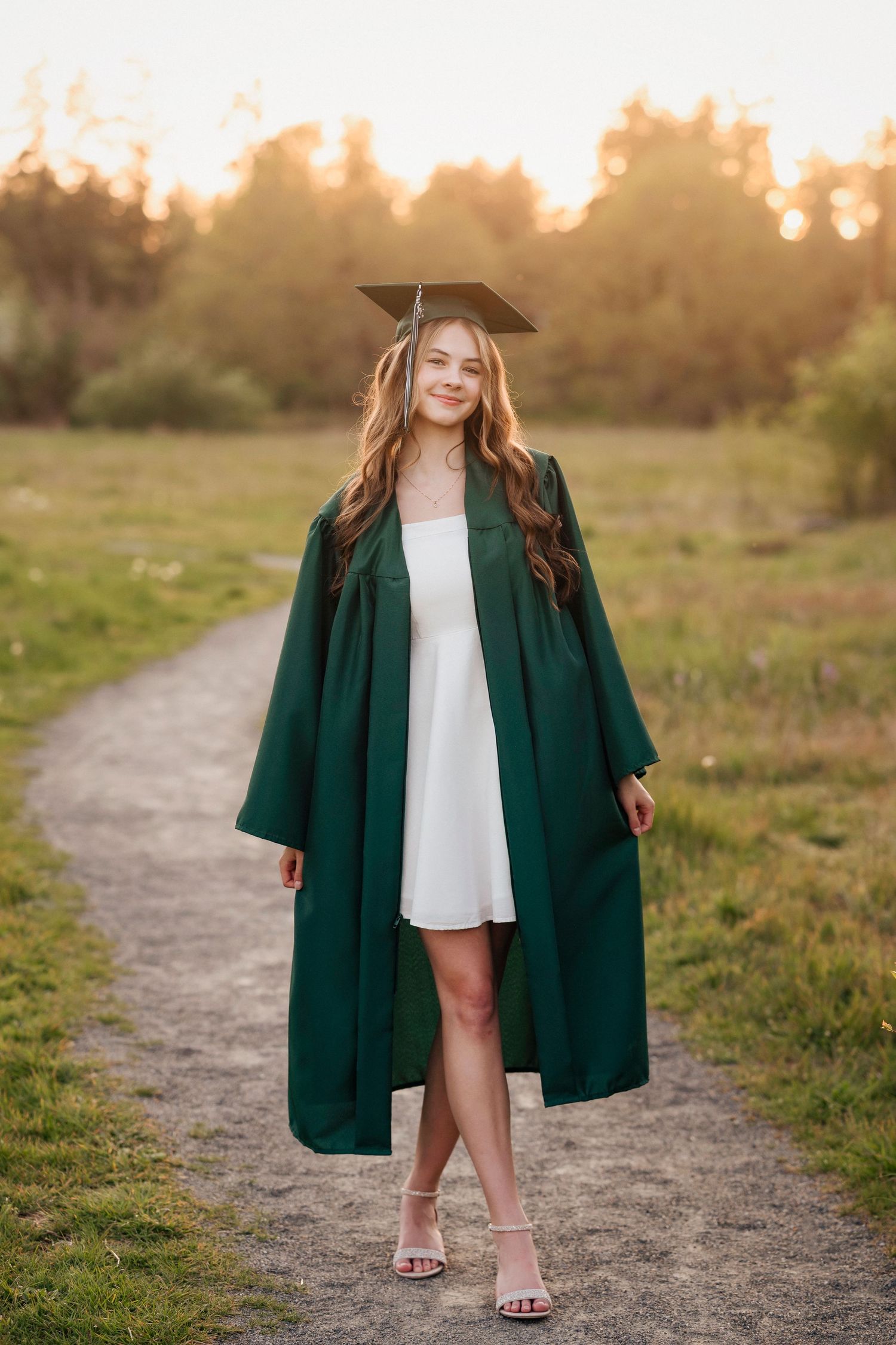 Graduate poses on nature trail wearing white dress and dark green graduation gown at sunset.