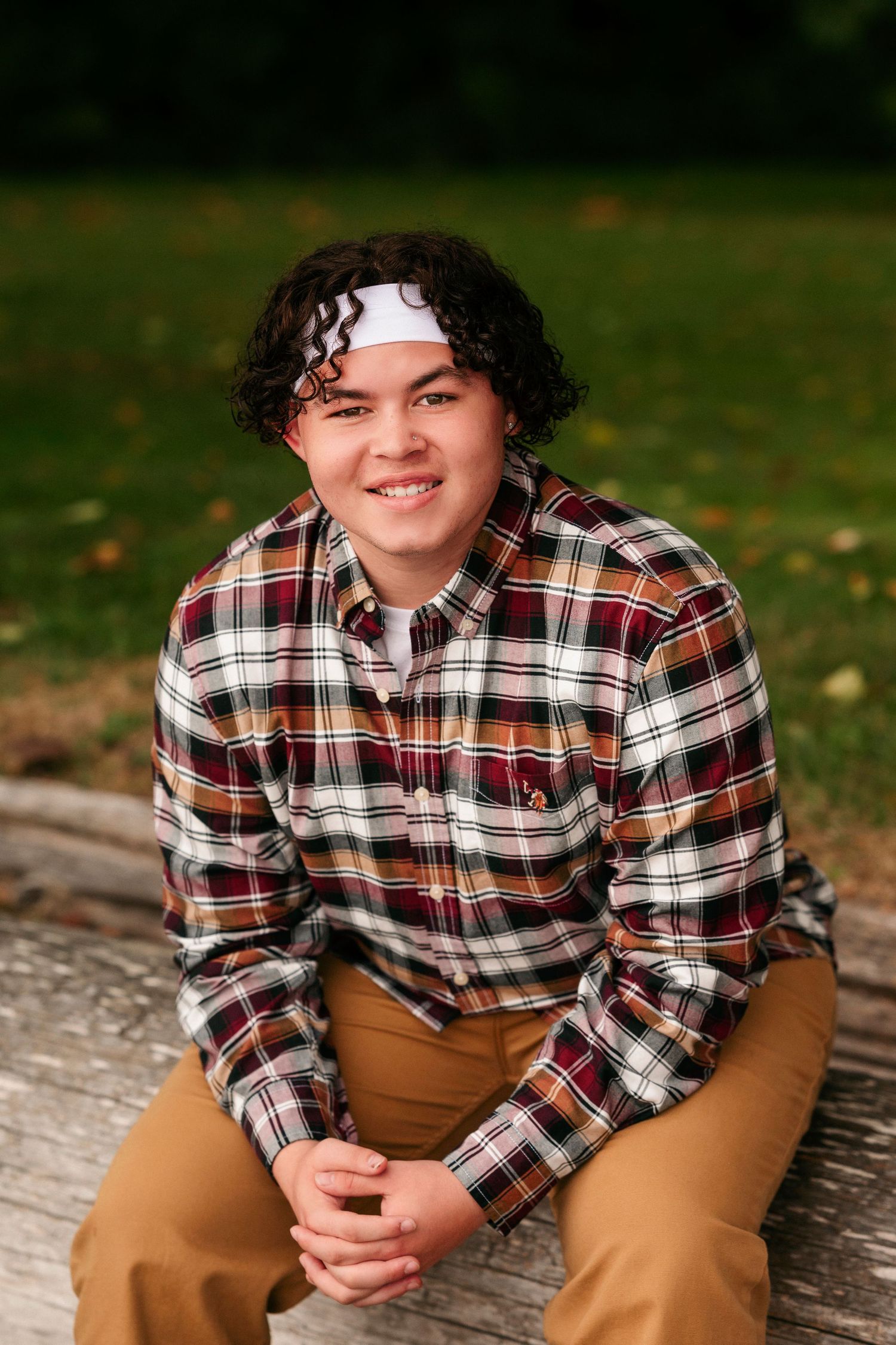 A person in a plaid flannel shirt and brown pants sits casually outdoors during autumn.