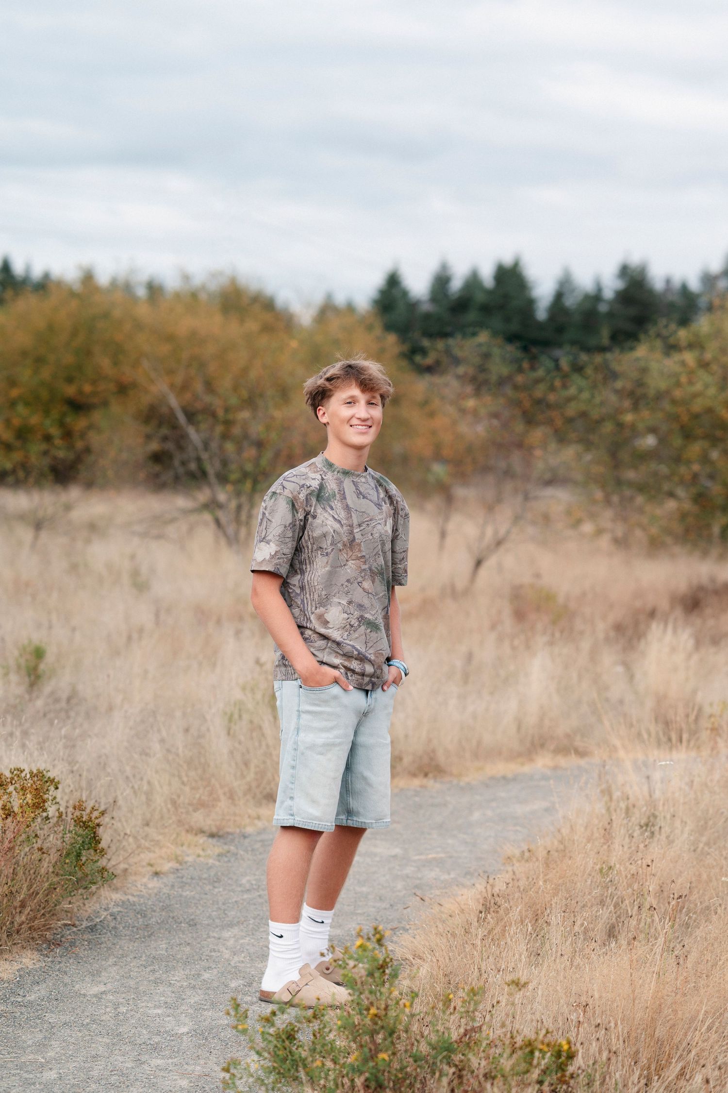 Someone in a camouflage t-shirt and jean shorts stands on a dirt path surrounded by dry grass and trees.