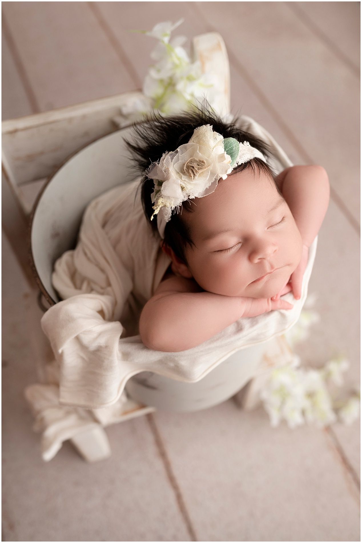 A sleeping newborn wearing a delicate floral headband lies peacefully on soft beige fabric surrounded by white flowers.