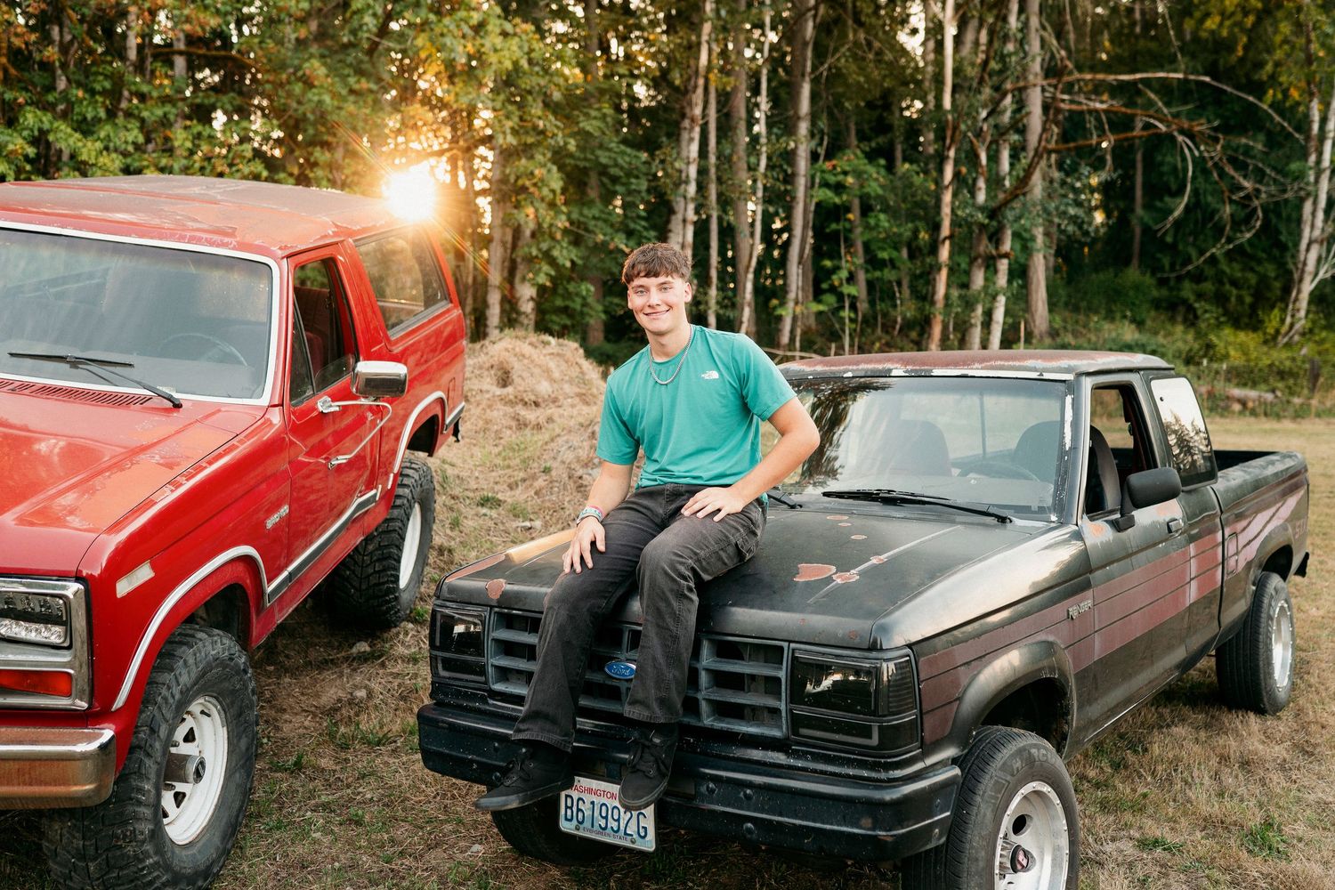 Two pickup trucks parked in a wooded area at sunset with a person leaning against one of the vehicles.