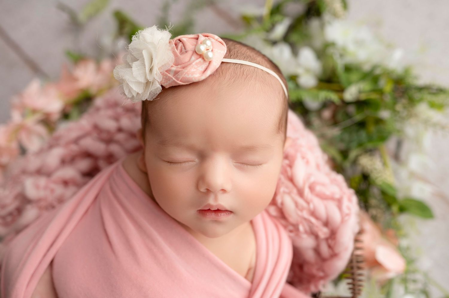 Sleeping baby wrapped in pink fabric wearing delicate floral headband with floral backdrop.