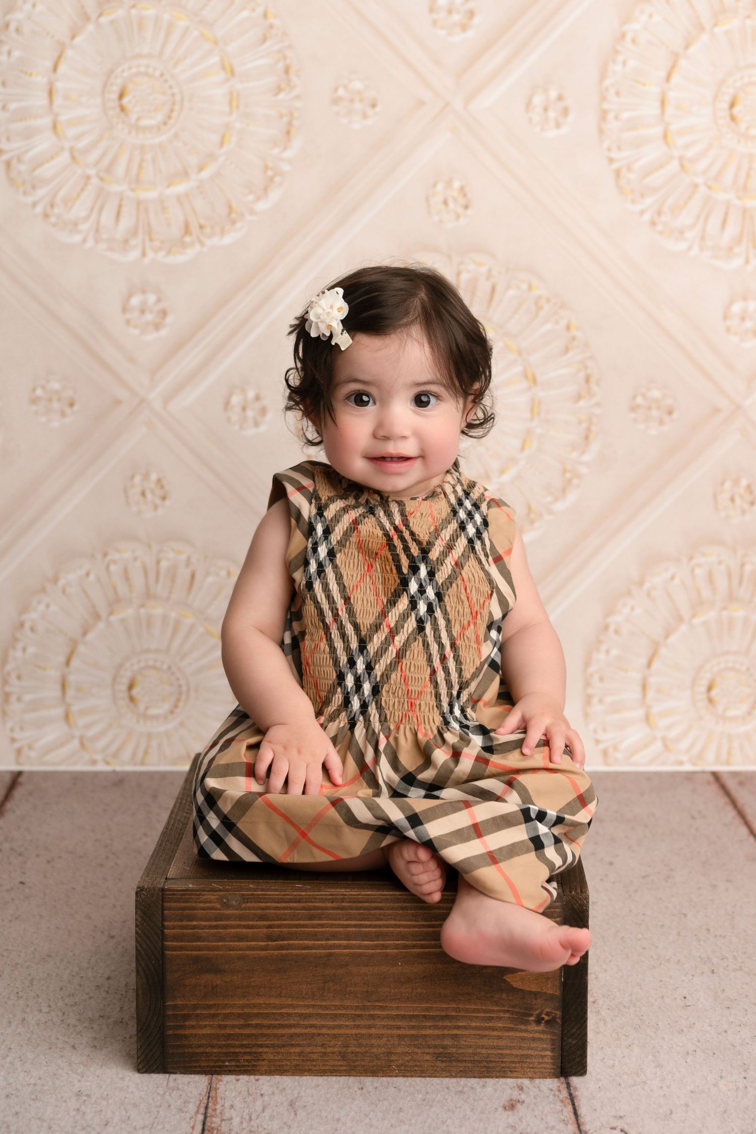 A small child in a plaid dress sits on a wooden box against a textured white wall.