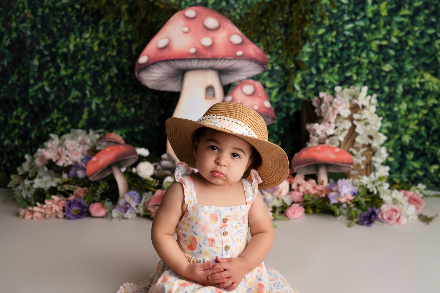 A child in a sun hat sits in front of a whimsical backdrop featuring large pink mushrooms and floral decorations.