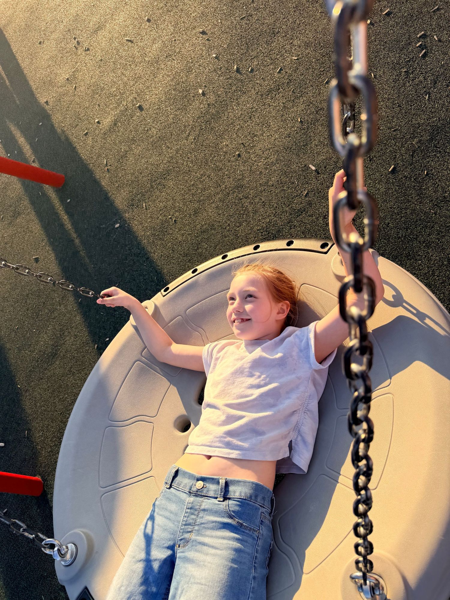 A smiling red-haired girl lies on a circular playground spinner, holding chains, wearing a white top and jeans.