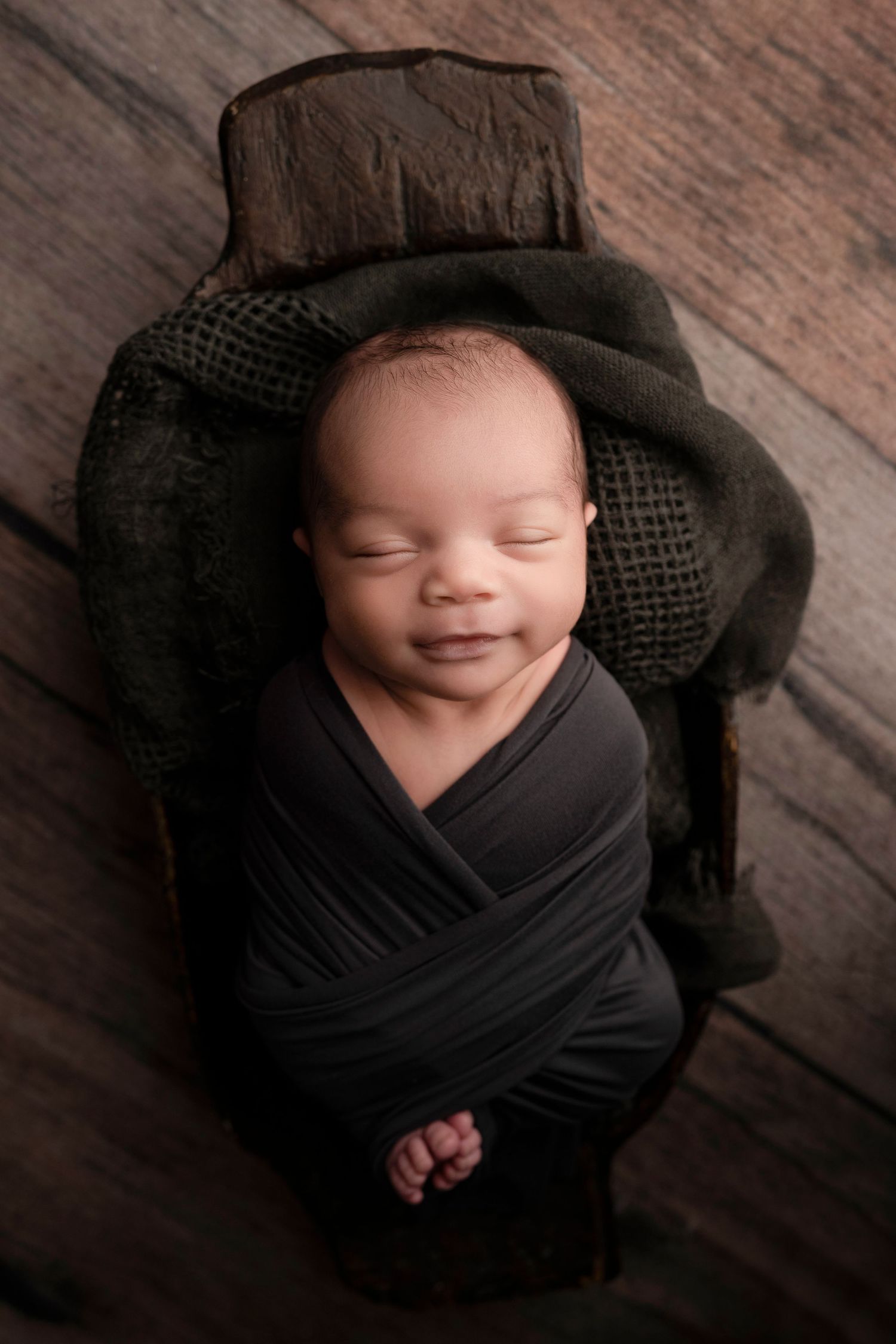 Smiling newborn baby swaddled in dark wrap, peacefully sleeping in a rustic wooden bowl on wood floor.