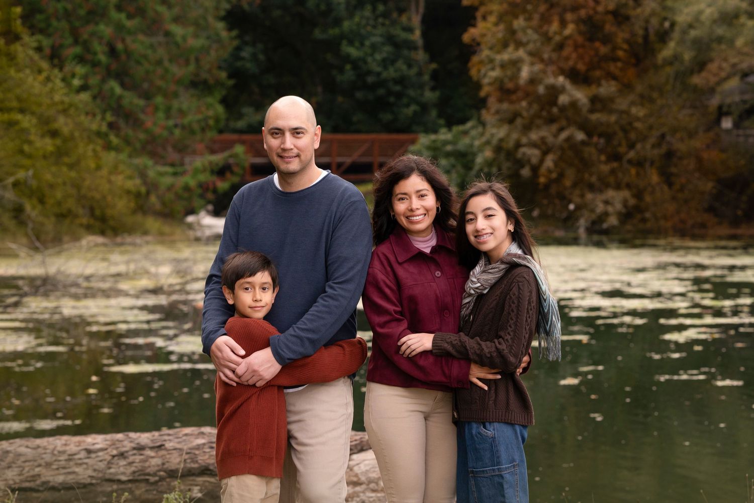 Family of four smiling outdoors near a serene pond with autumn foliage in the background.