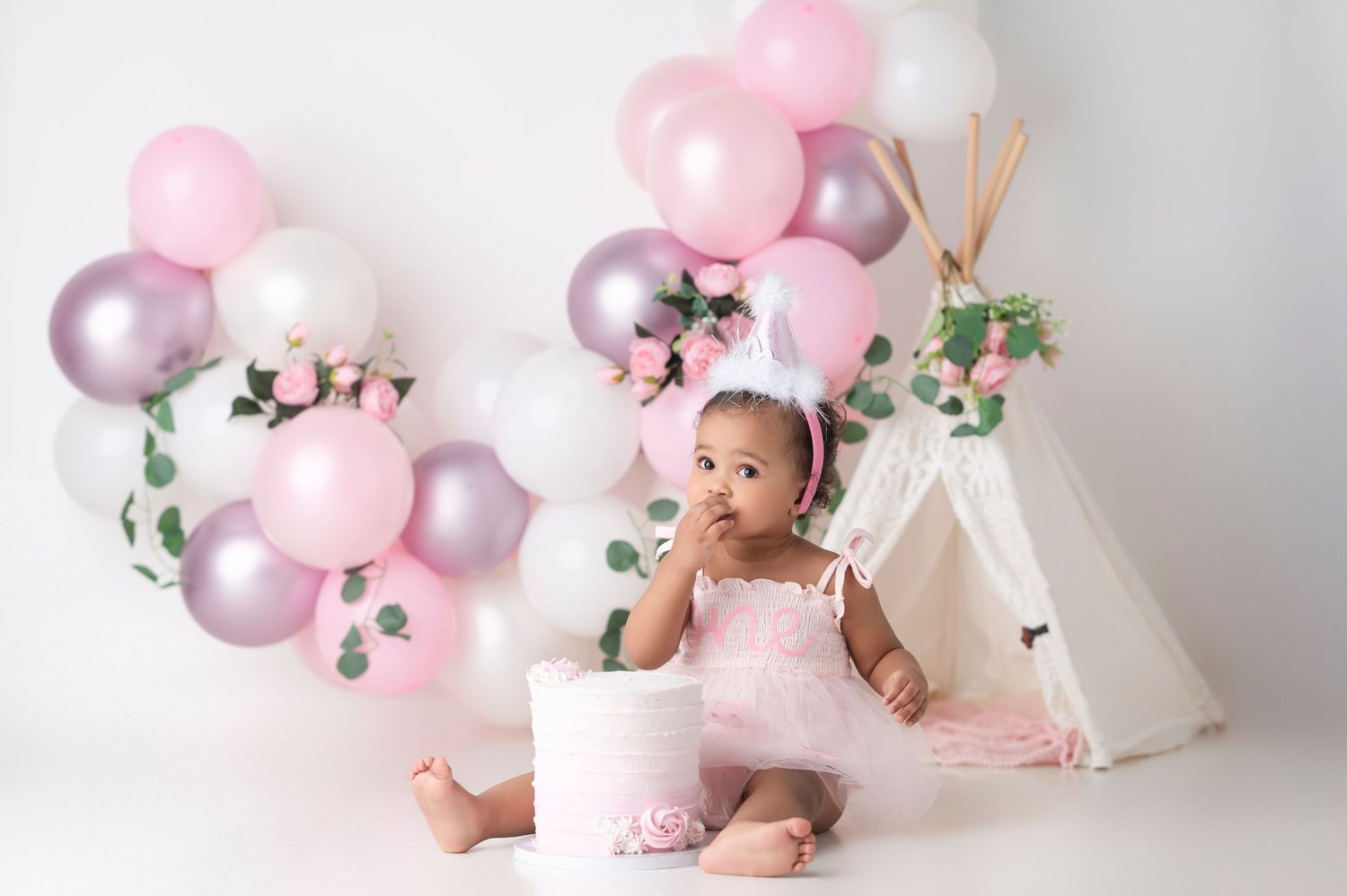 Baby girl in pink tutu sits before a white teepee with pink, white, and purple balloons for her first birthday cake smash.