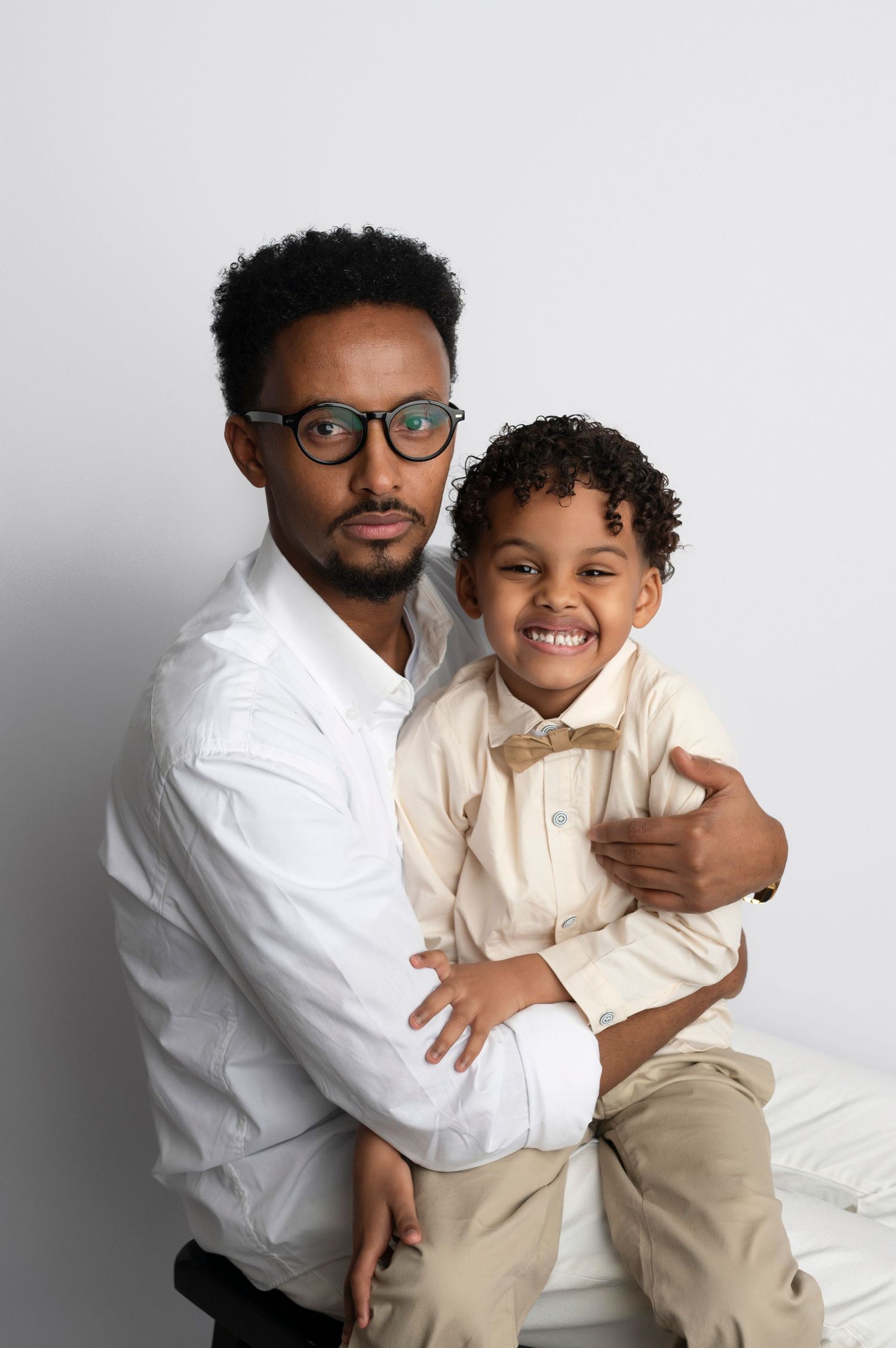 A man wearing glasses hugs a smiling young boy in a bow tie, both dressed in light-colored shirts for a portrait.