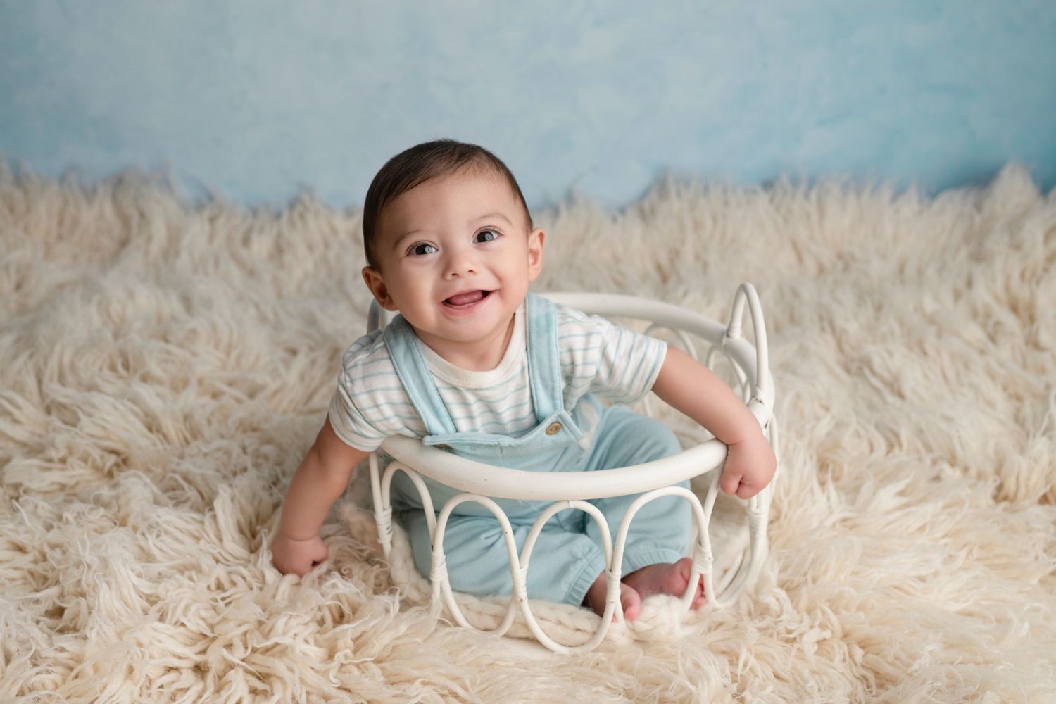 Smiling baby boy in blue romper sitting in white wicker basket on fluffy cream rug with light blue backdrop.