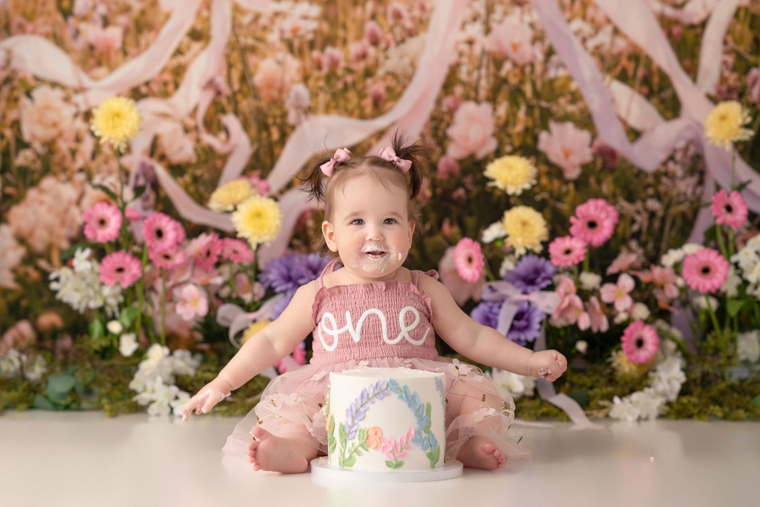 Baby girl in pink tutu sits before a floral backdrop with a white “one” cake for her first birthday cake smash.