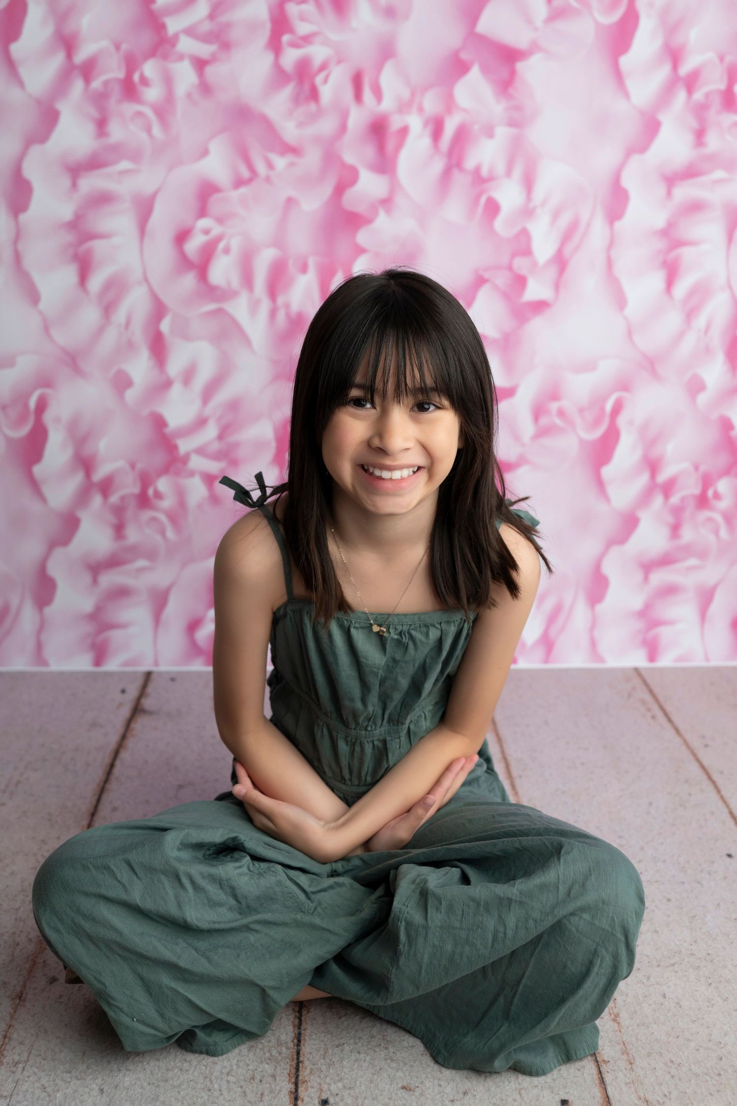 A smiling girl in a green jumpsuit sits cross-legged on a wooden floor against a pink floral backdrop.