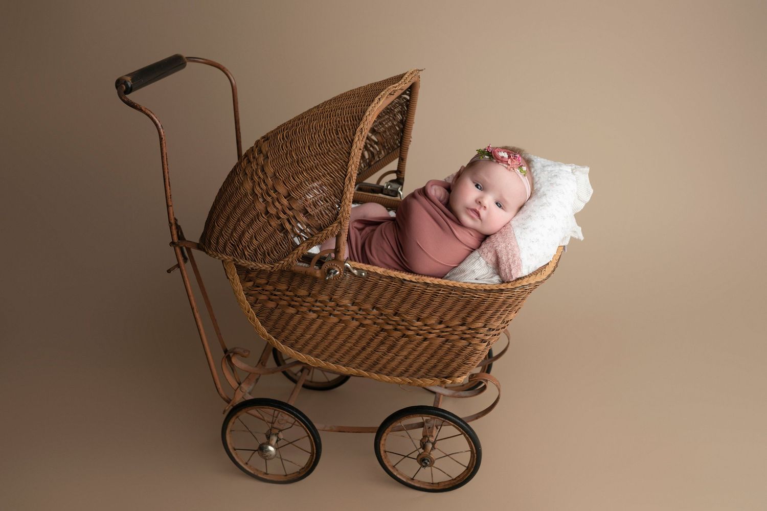 Newborn baby sleeping peacefully in a vintage wicker pram, wrapped in pink, with a white pillow on a tan background.
