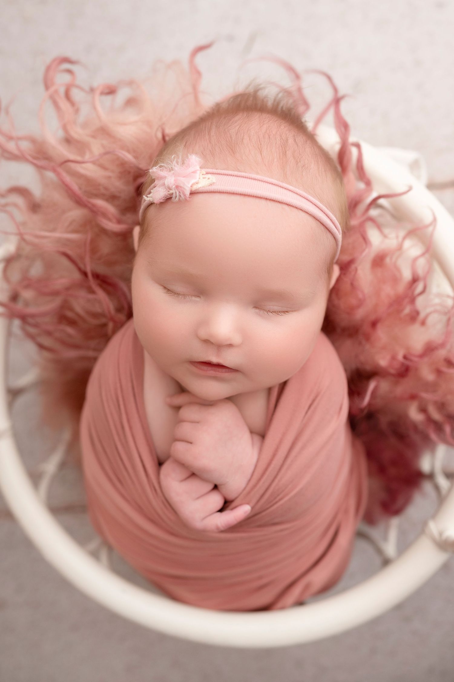 Newborn baby girl swaddled in pink wrap, sleeping peacefully in white basket with pink fluffy fur and floral headband.