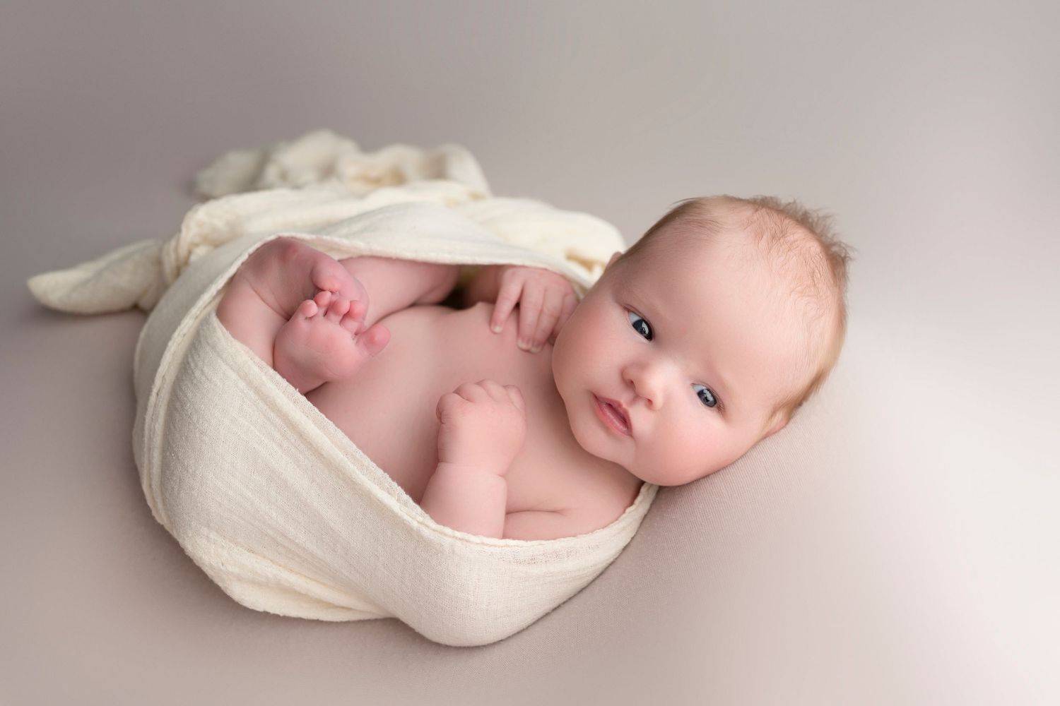 Newborn baby wrapped in cream fabric, lying on gray background in professional newborn photography portrait.