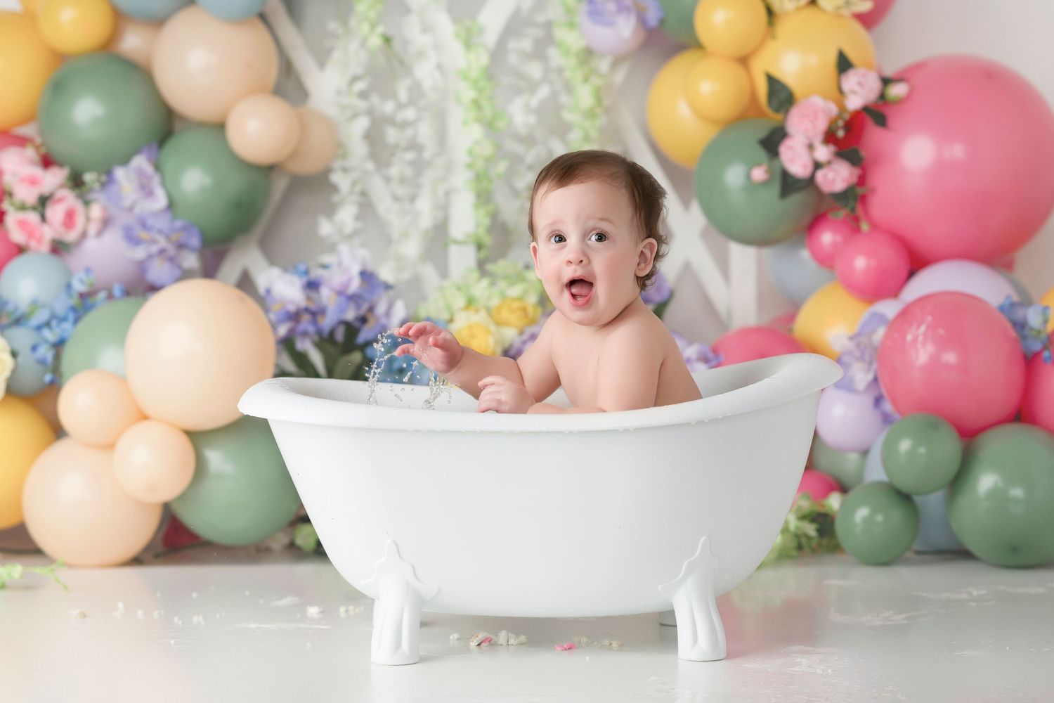 A baby smashes into a white frosted cake while colorful balloons decorate the background in a cake smash photo session.