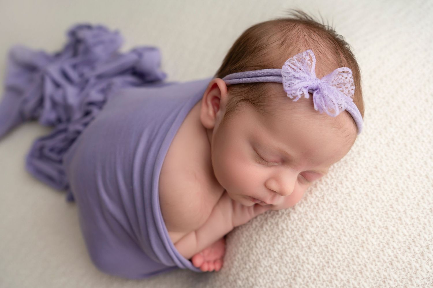 A sleeping baby wearing a purple outfit and lavender bow headband rests peacefully on a white surface.