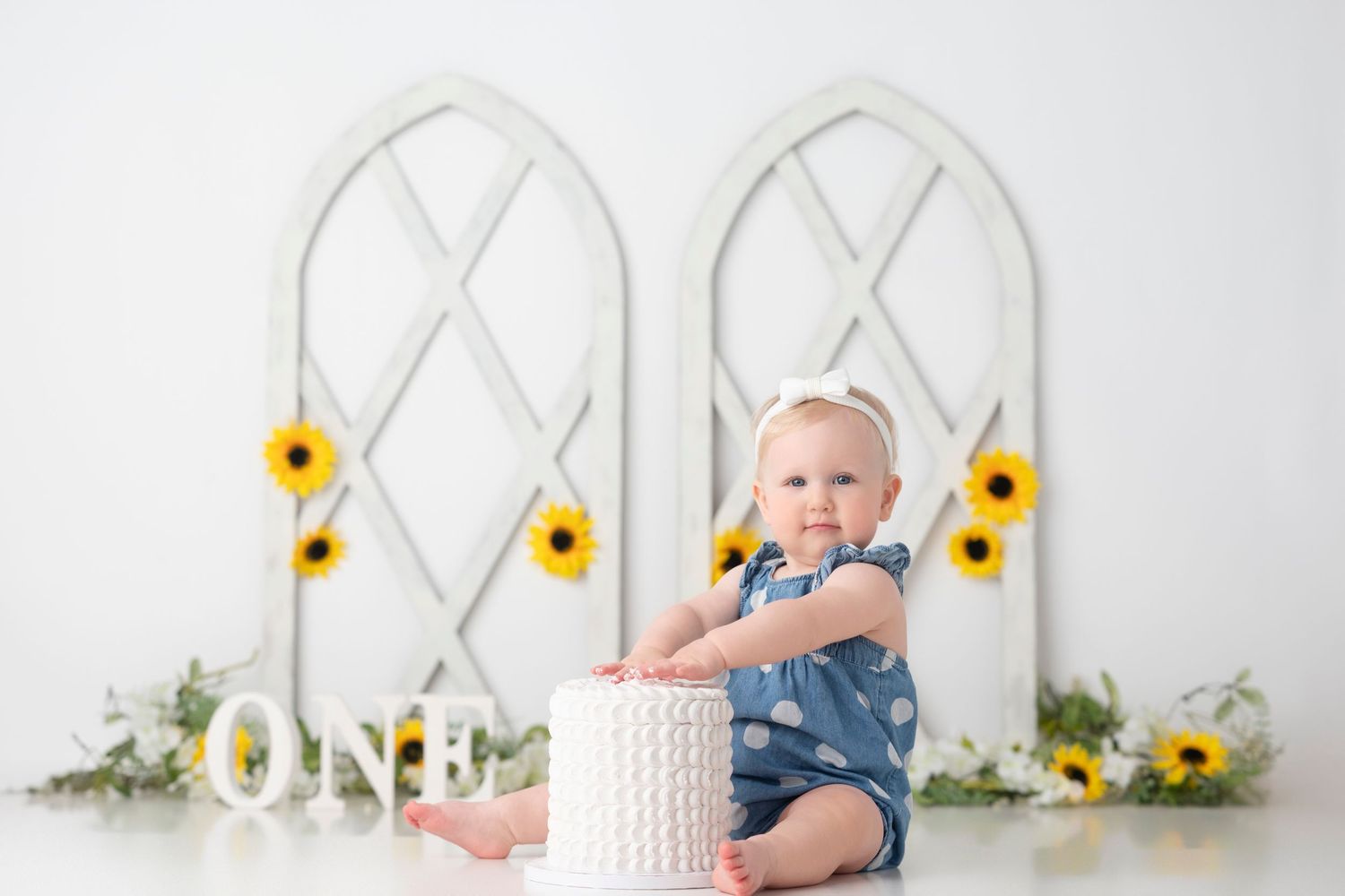 Baby in denim romper sits on white floor with sunflower and trellis backdrop during milestone photo session.