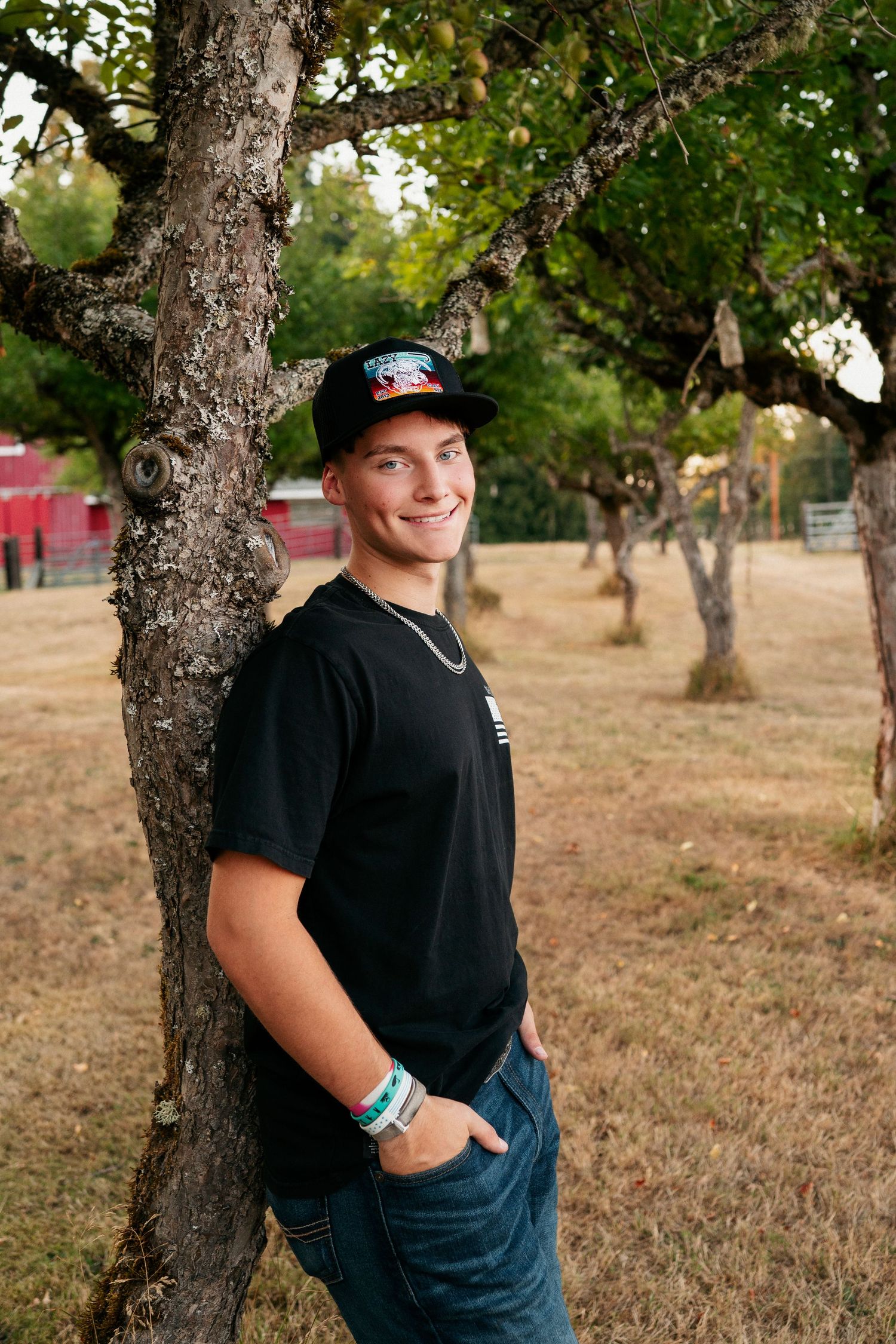 Person wearing a black shirt and baseball cap poses casually against a tree trunk in a grassy outdoor setting.