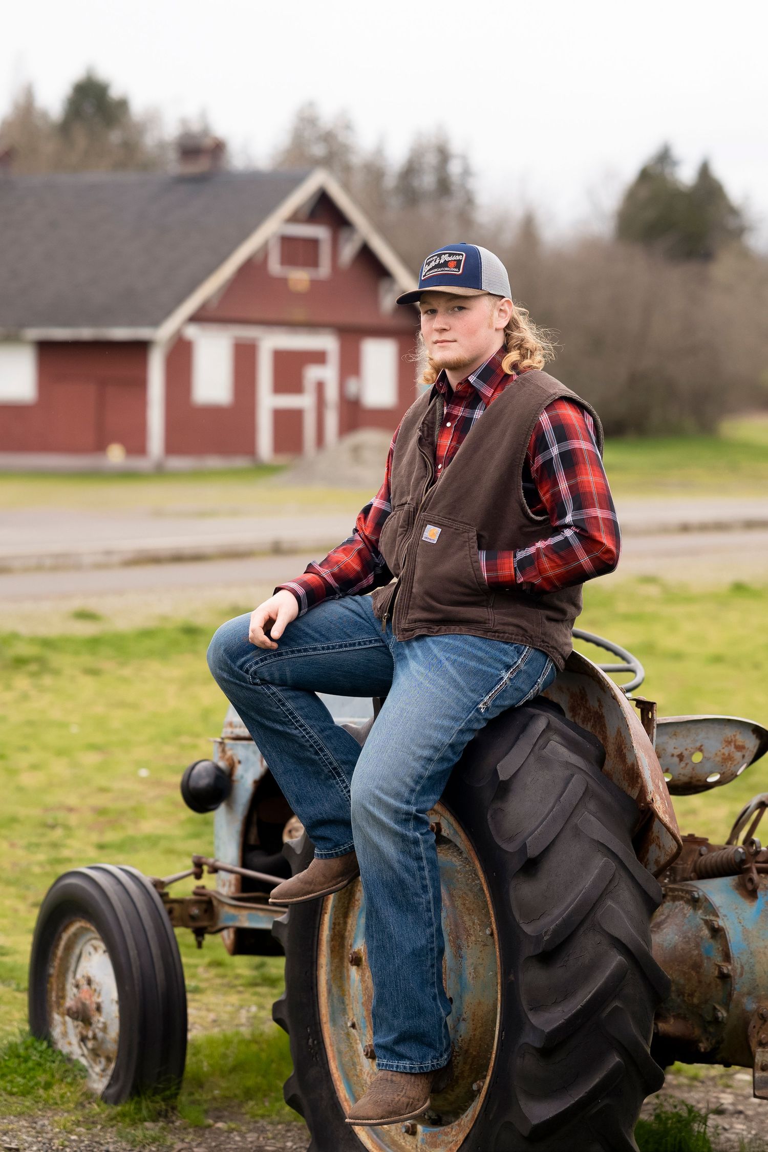A rustic farm scene showing someone sitting on an old tractor in a plaid shirt and work vest against red barns.