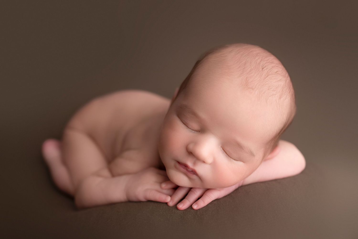 Sleeping newborn baby curled up peacefully against brown backdrop in artistic portrait style.
