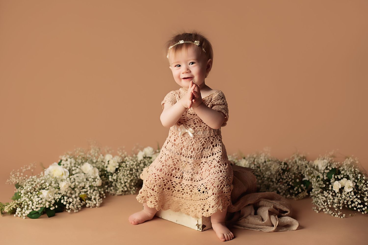 A small child in a lace dress kneels on a brown backdrop surrounded by delicate white floral arrangements.